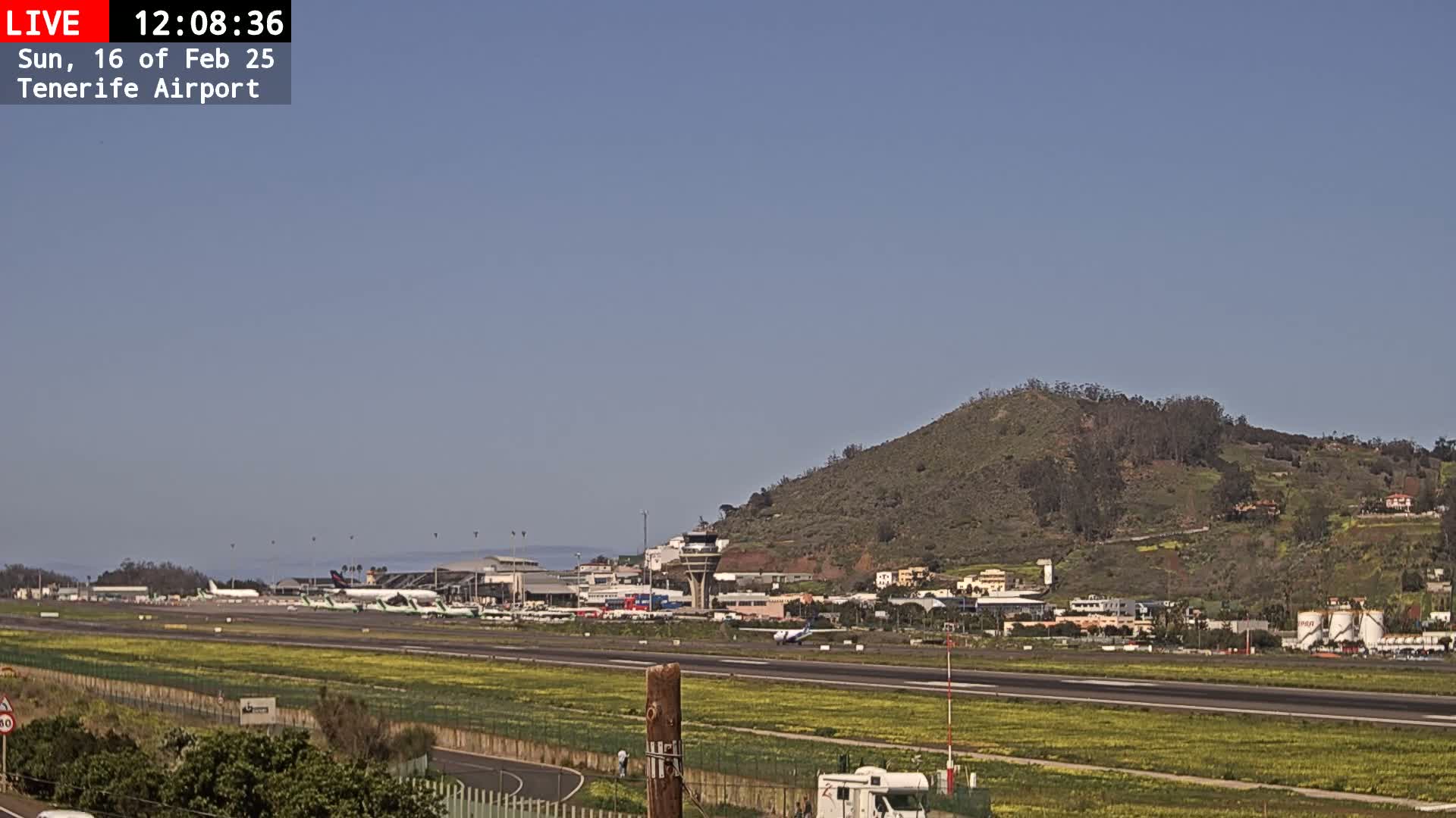 A sunny day at an airport shows several airplanes on the tarmac, with a control tower and a hill in the background.