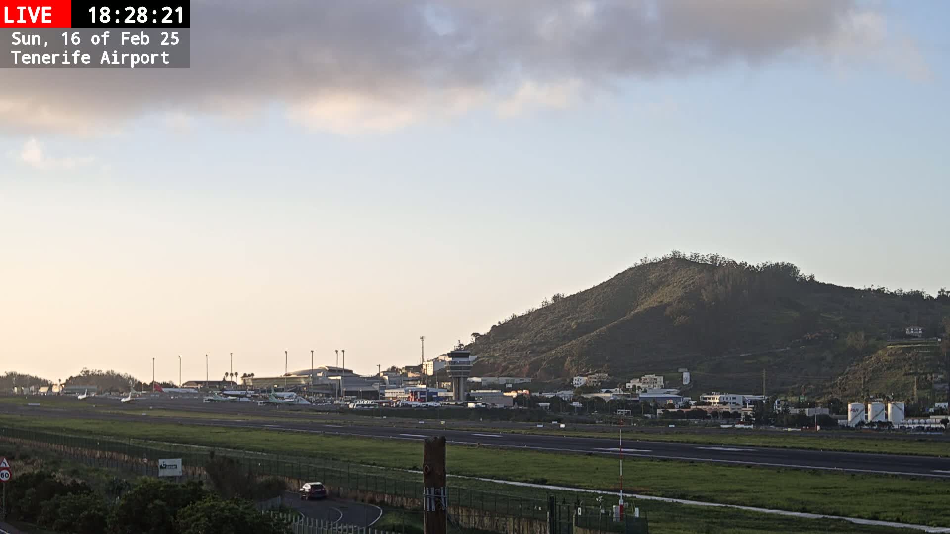 A mostly cloudy sunset illuminates an airport runway and terminal buildings situated beneath a hill.