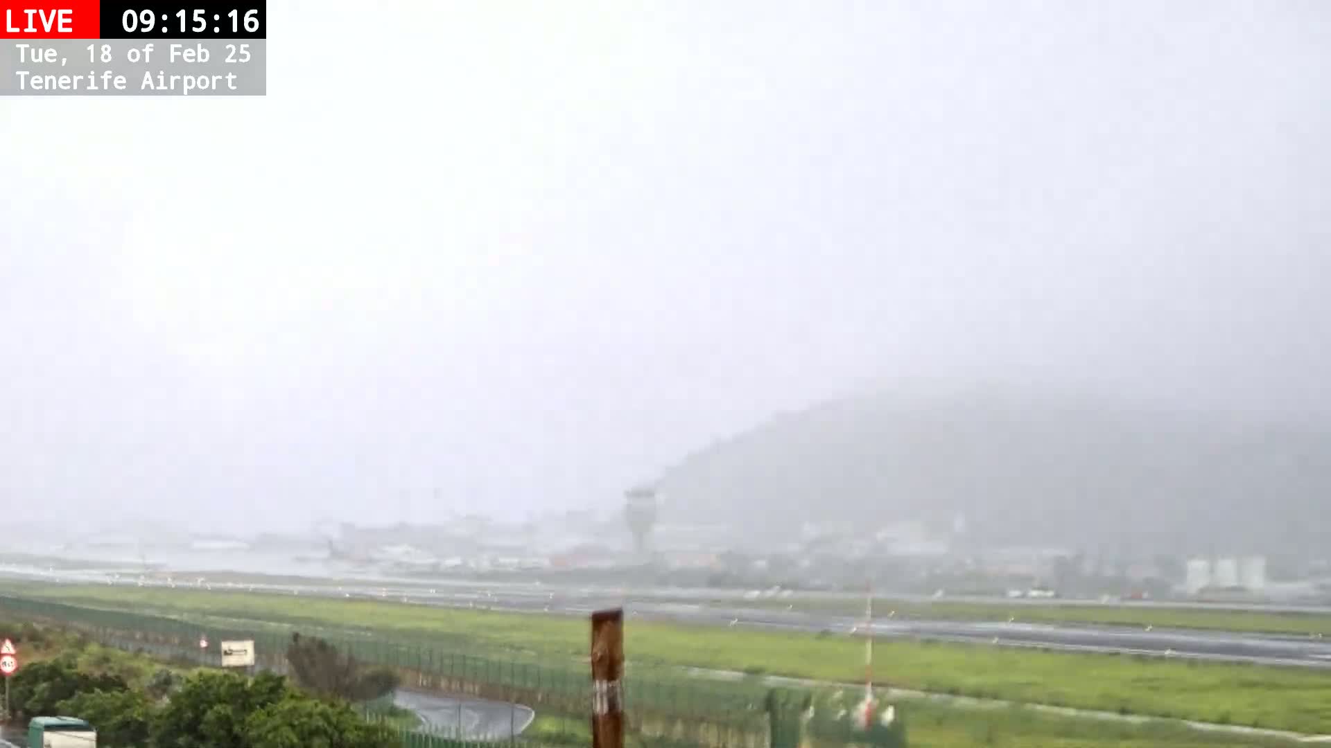 A rain-covered view of an airport runway and surrounding area, with a hazy mountain visible in the background.