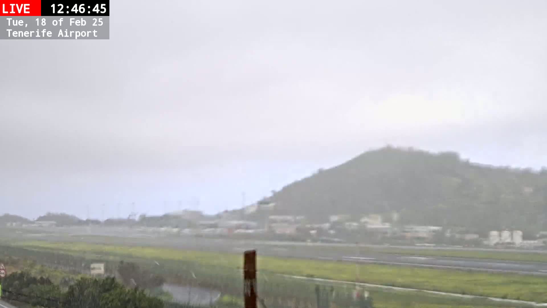 A rainy view of a flat, grassy area near a runway, with a hill visible in the background under an overcast sky.