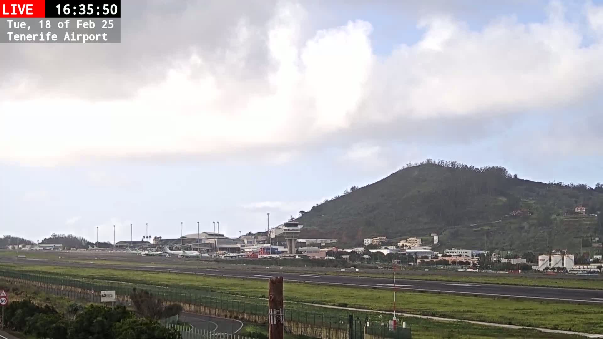 An airport runway with several planes parked, surrounded by grassy areas and a hill in the background under a partly cloudy sky.