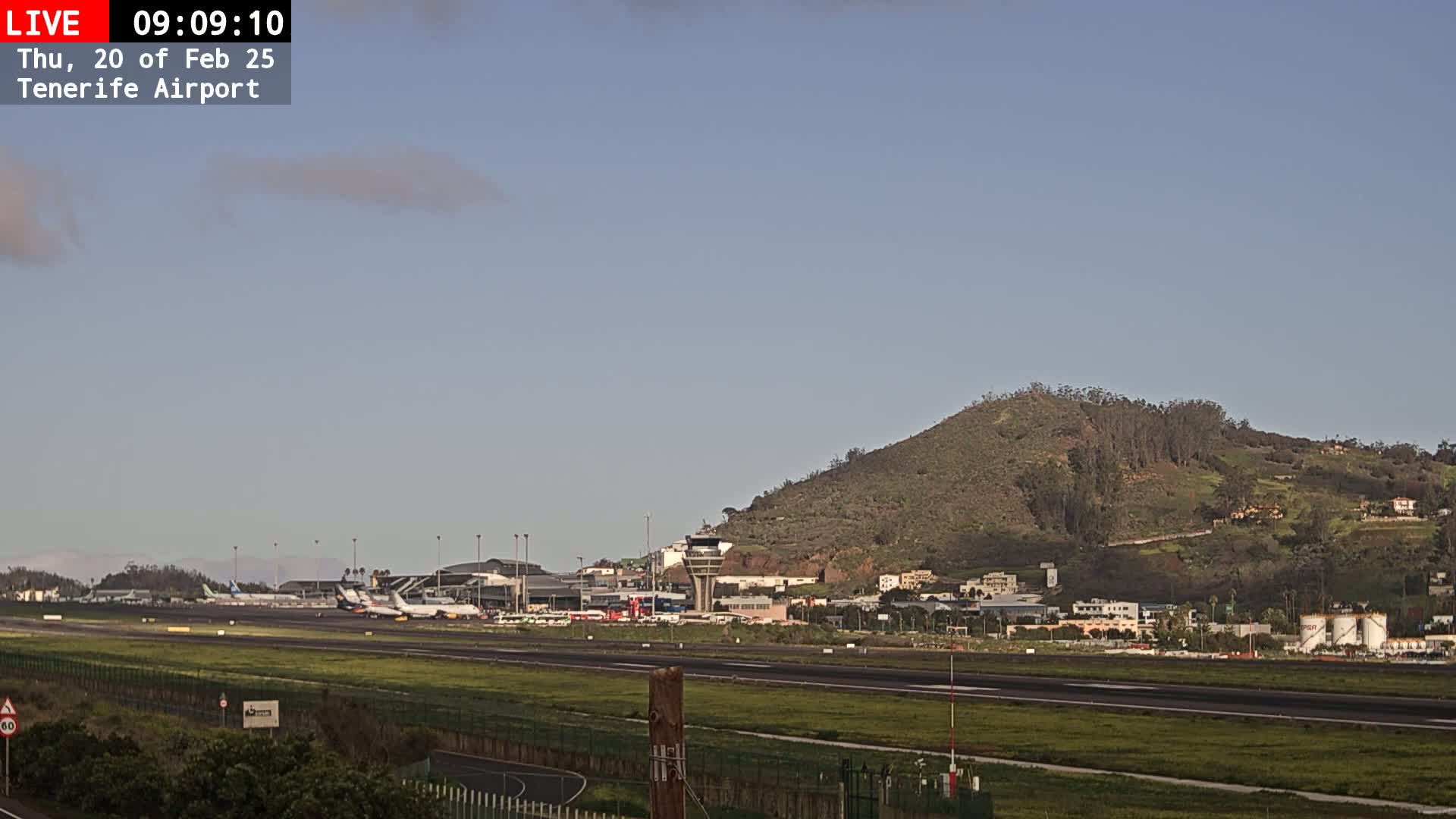 A mostly clear sky overlooks an airport with several airplanes on the tarmac, a control tower, and a hill in the background.