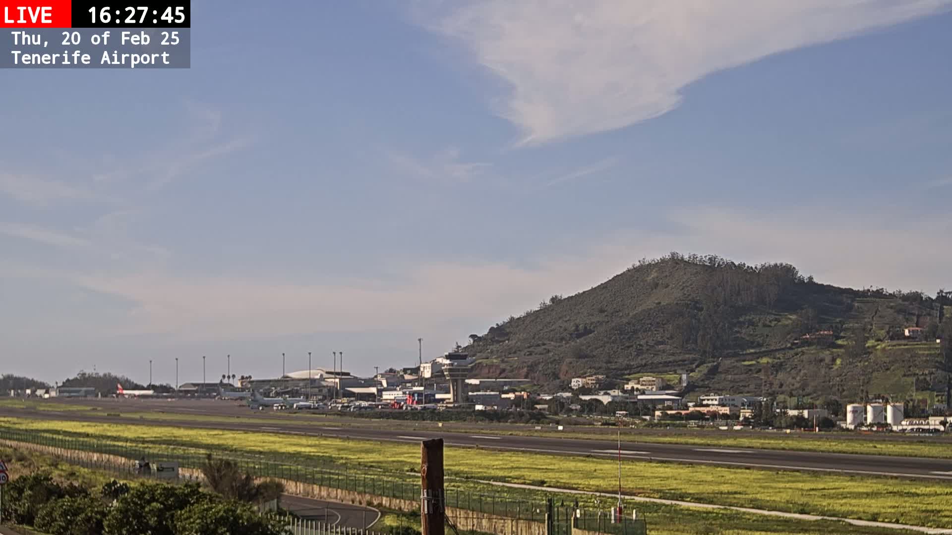 A view of an airport runway, with several airplanes visible, set against a partly cloudy sky and a brown, vegetated hill in the background.