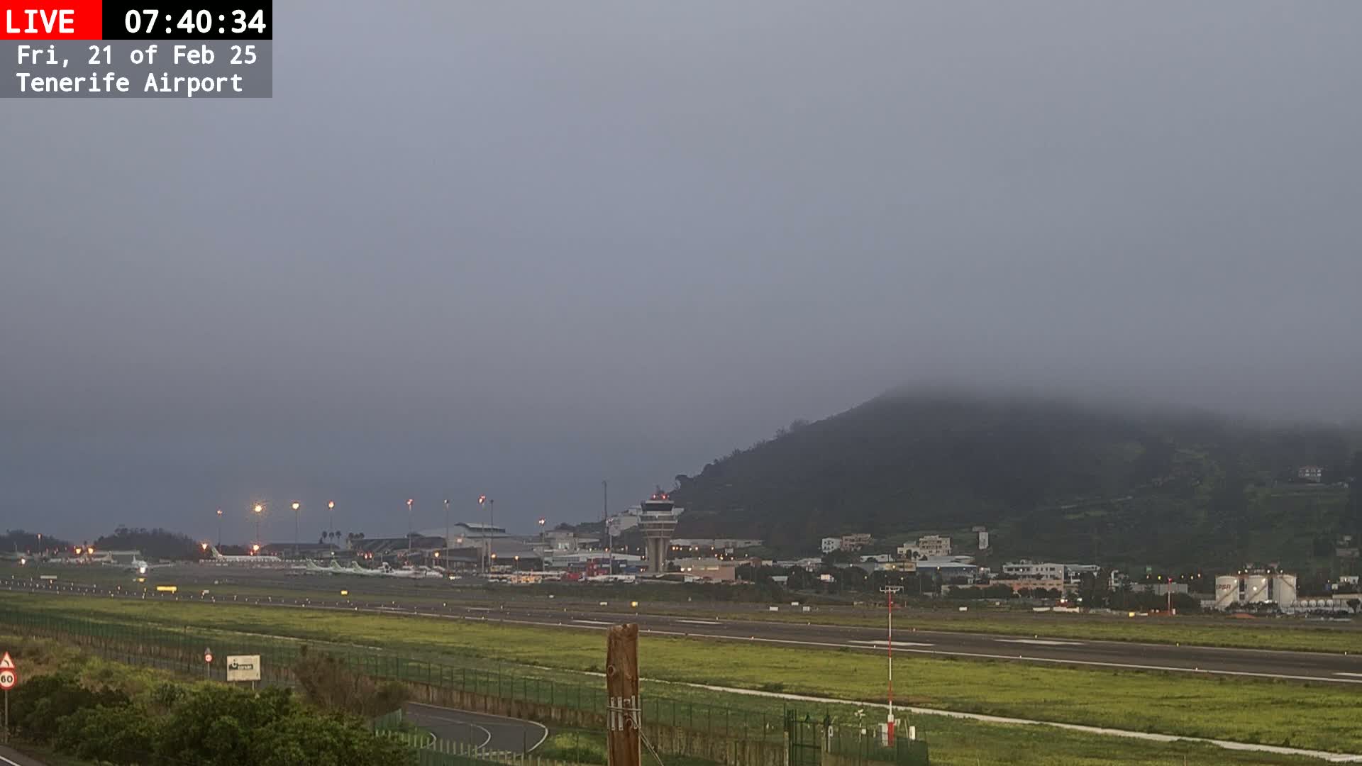 A foggy sunrise view of an airport runway, with several airplanes parked on the tarmac and a hill shrouded in mist in the background.