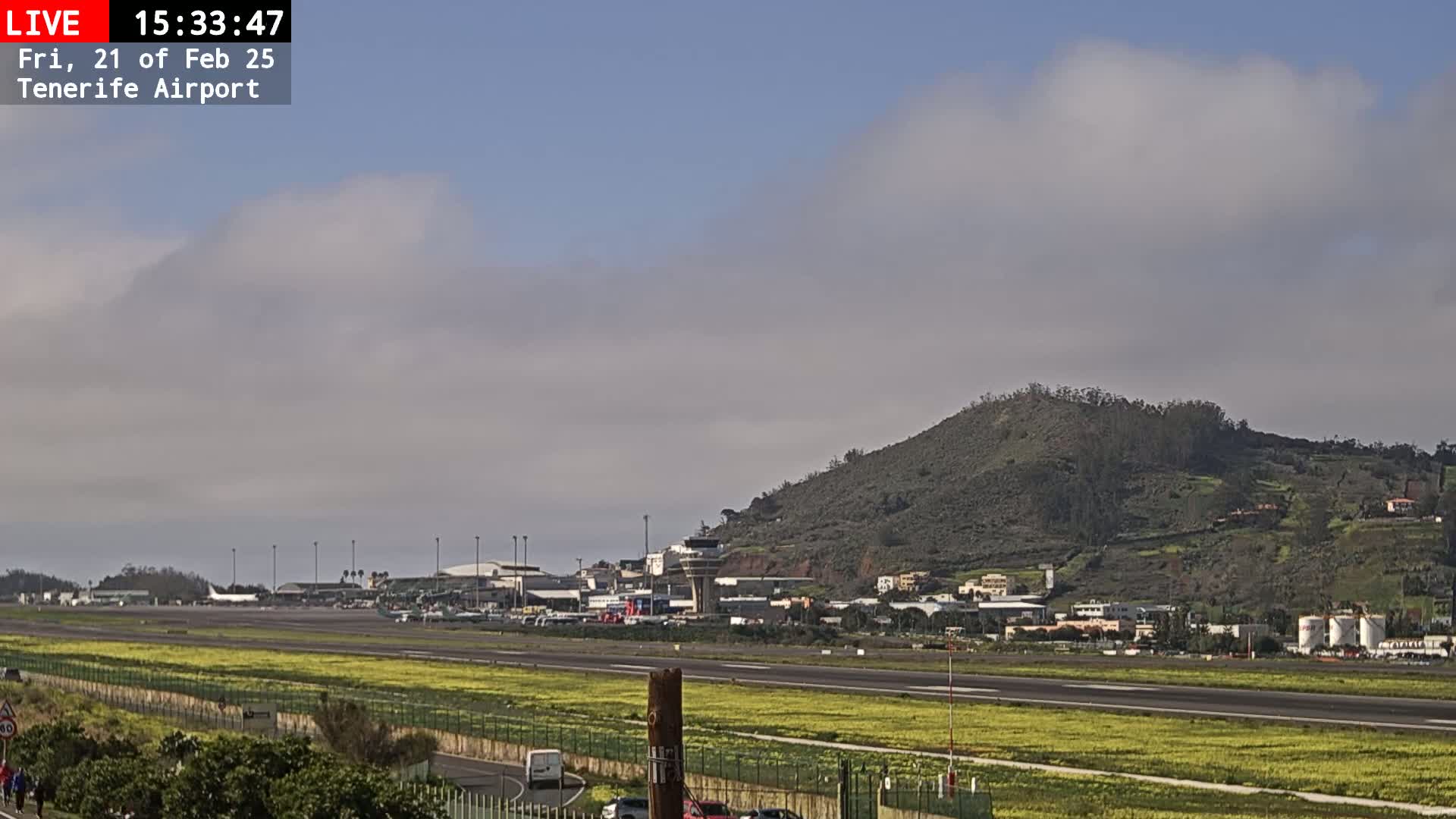 A view of an airport runway with several planes visible, a control tower, and a large hill in the background under a partly cloudy sky.
