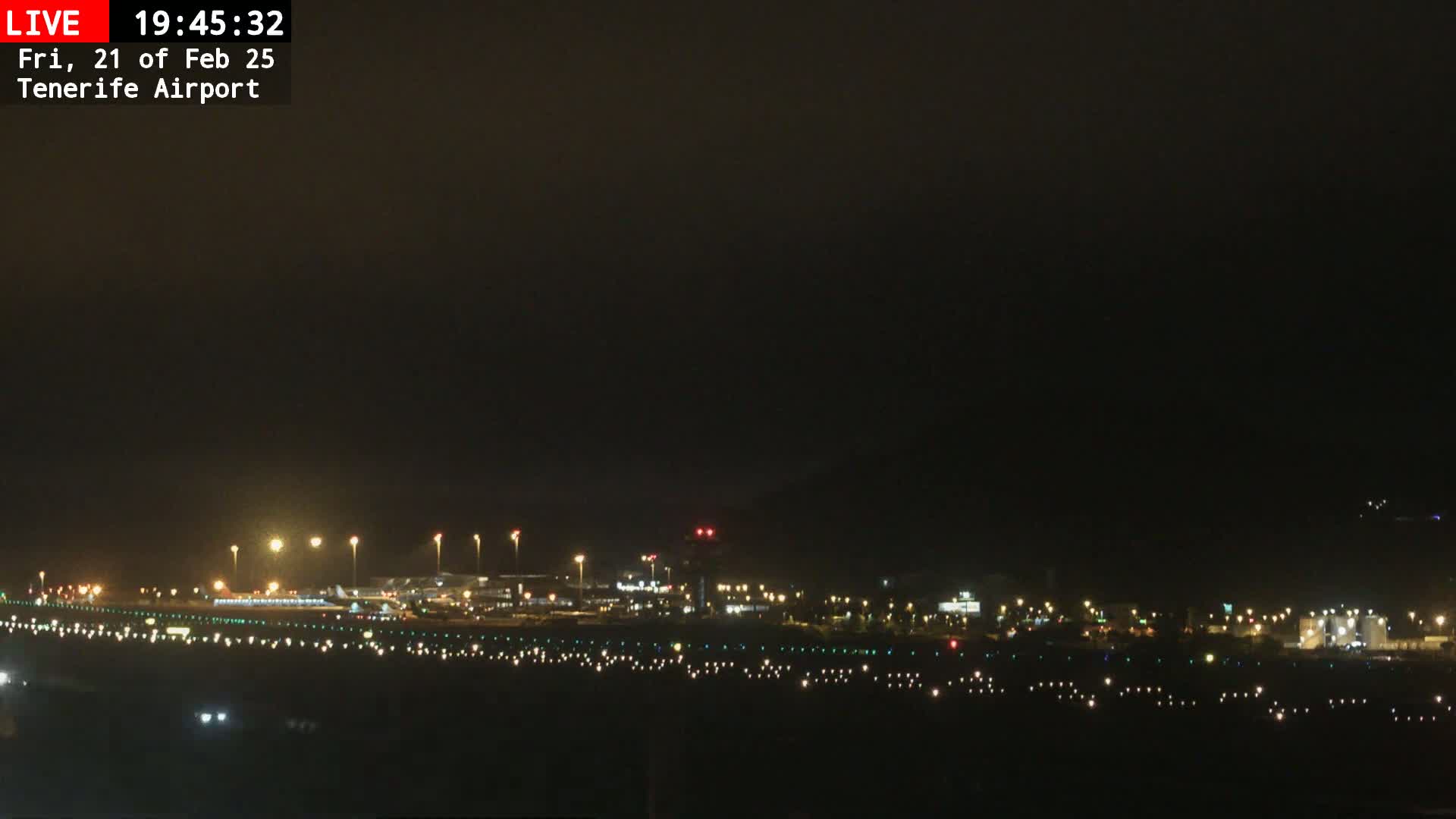 A nighttime view of an airport runway and terminal buildings illuminated by lights under a dark, overcast sky.