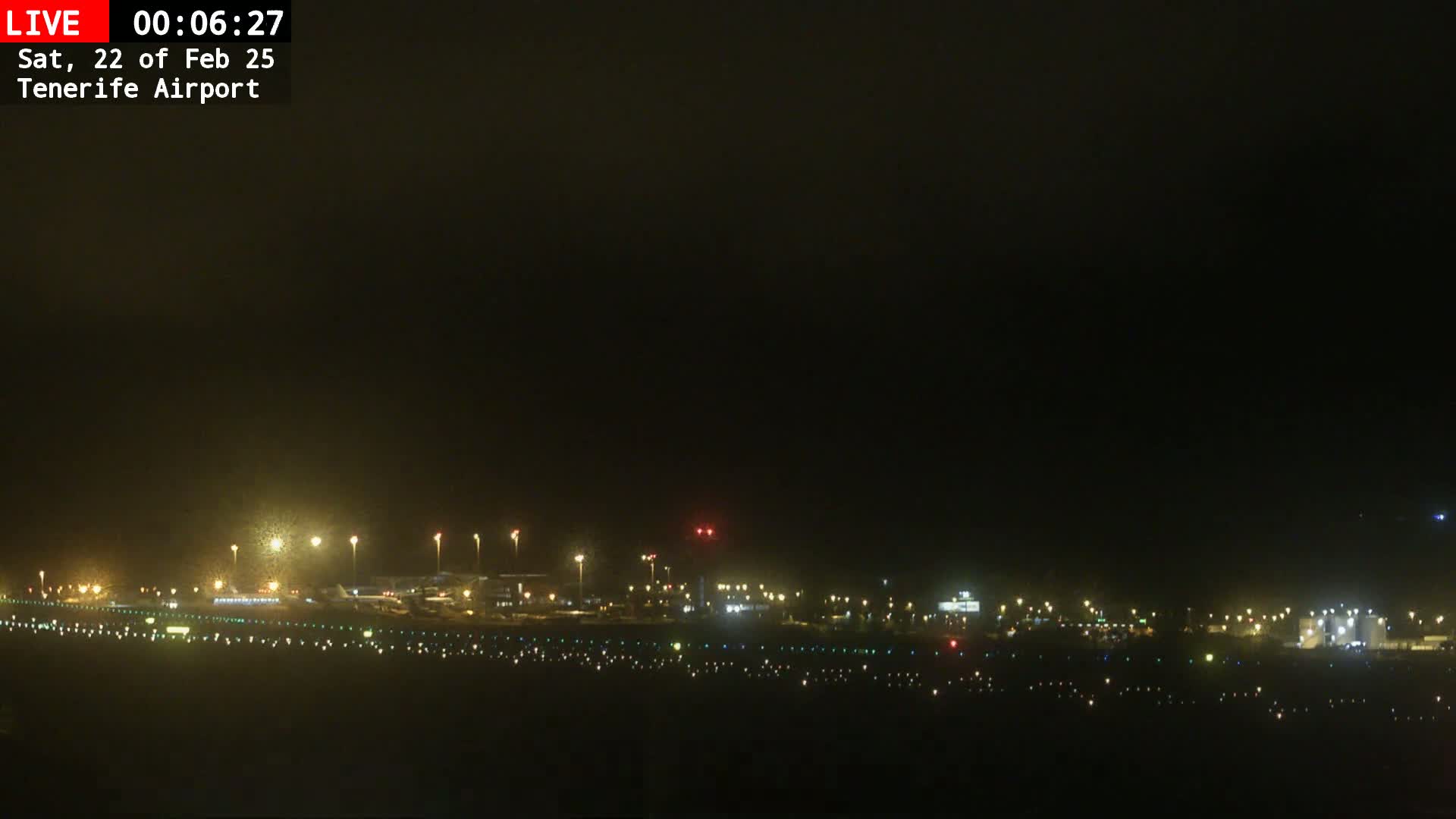 A nighttime view of an airport runway and terminal buildings under a dark, hazy sky.