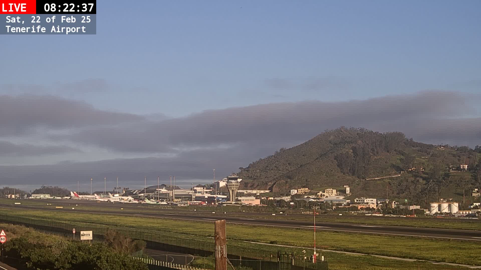 A view of an airport runway with several airplanes parked, a control tower, and a hill in the background under a partly cloudy sky.
