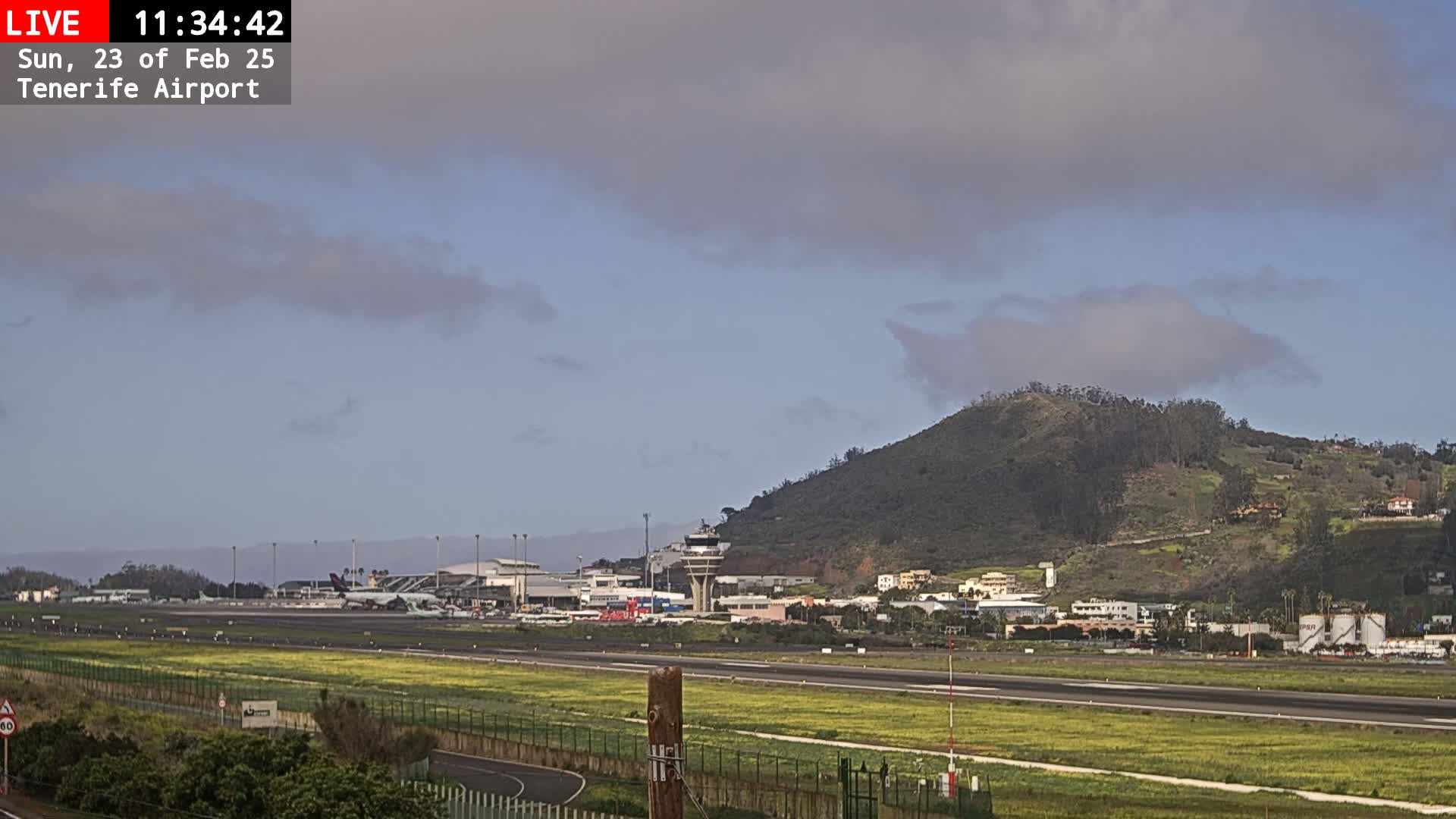 An airport runway and terminal are visible beneath a partly cloudy sky, with a green hill in the background.