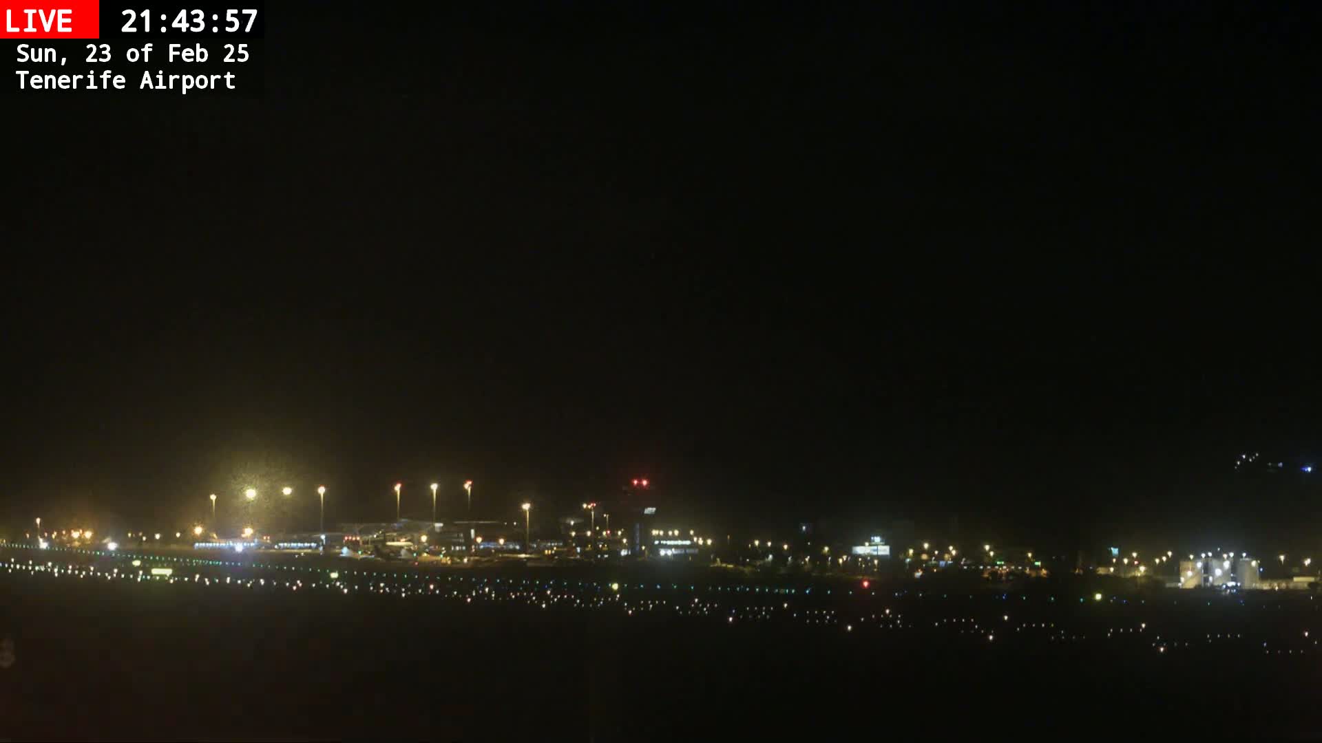 A nighttime long shot of a brightly lit airport runway and terminal buildings under a clear, dark sky.