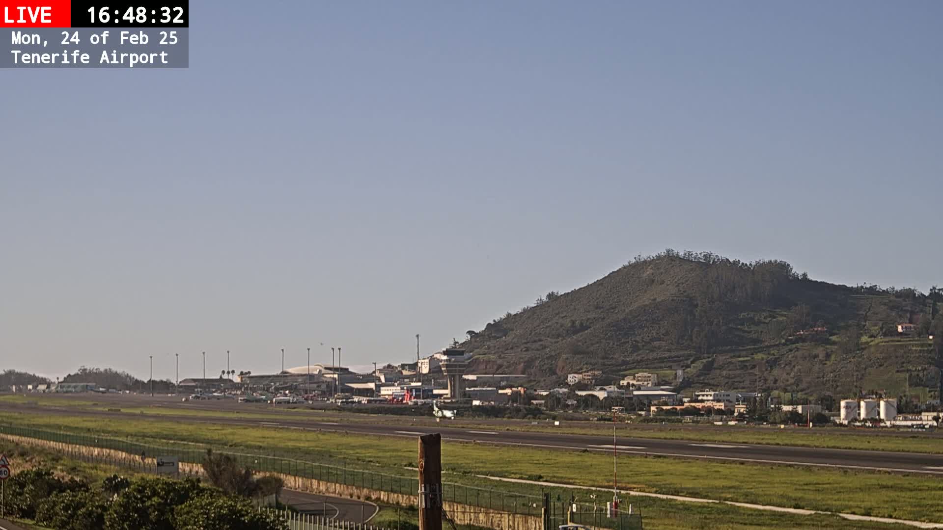 A long shot of an airport runway, terminal buildings, and a large hill in the background under a clear, bright sky.