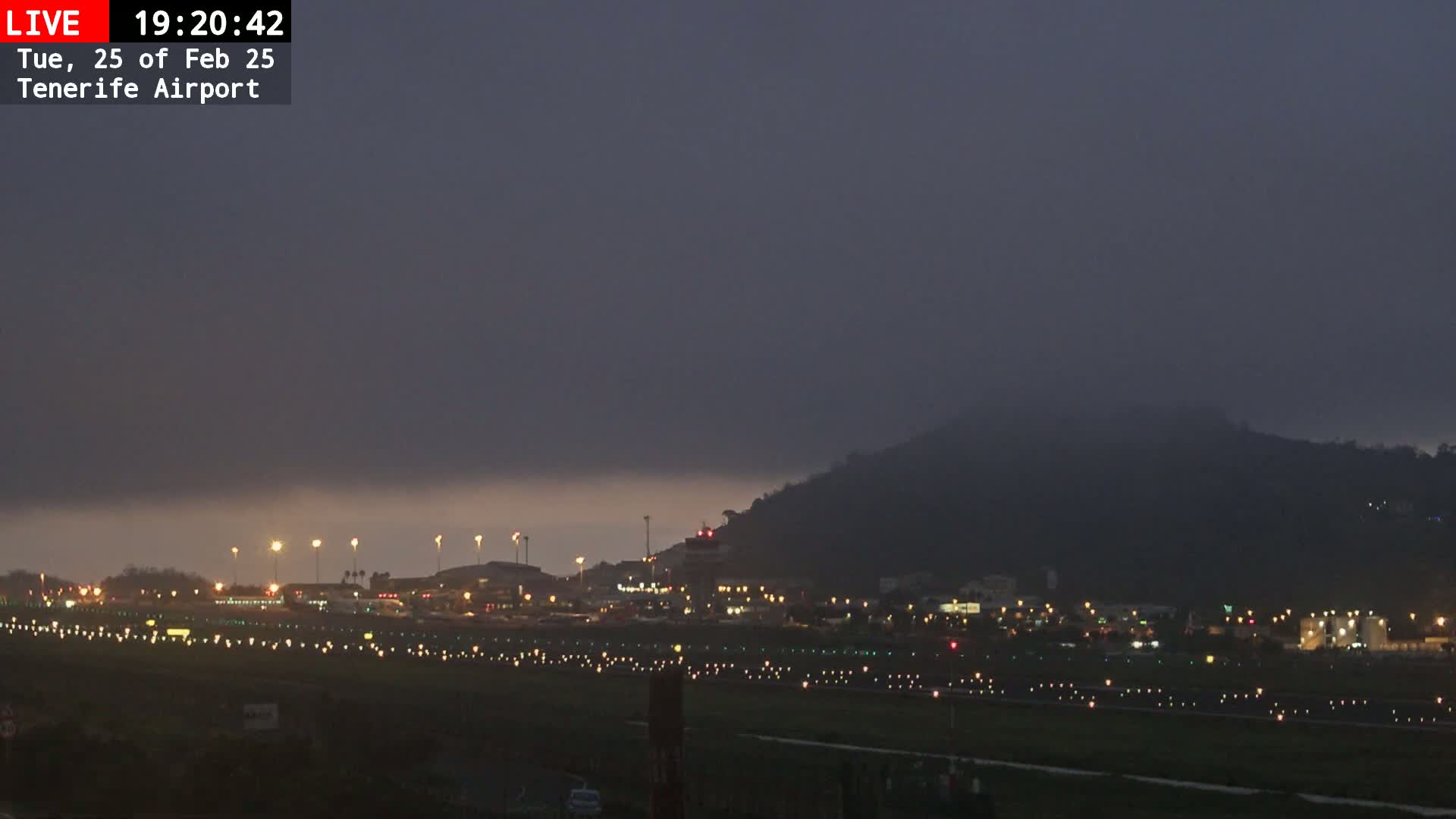 A low-lying fog obscures a hill overlooking a brightly lit airport at twilight.