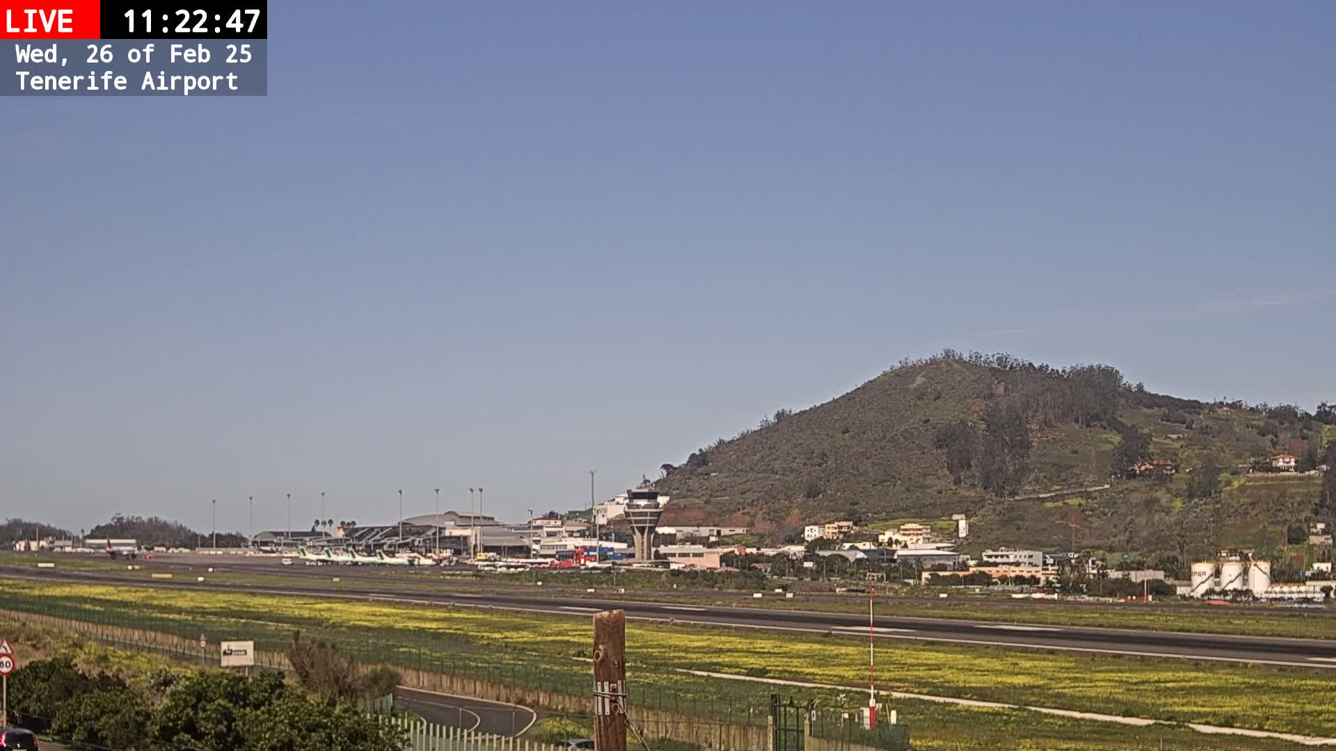 A sunny day at an airport shows several airplanes parked on the tarmac, with a control tower and a hill in the background.
