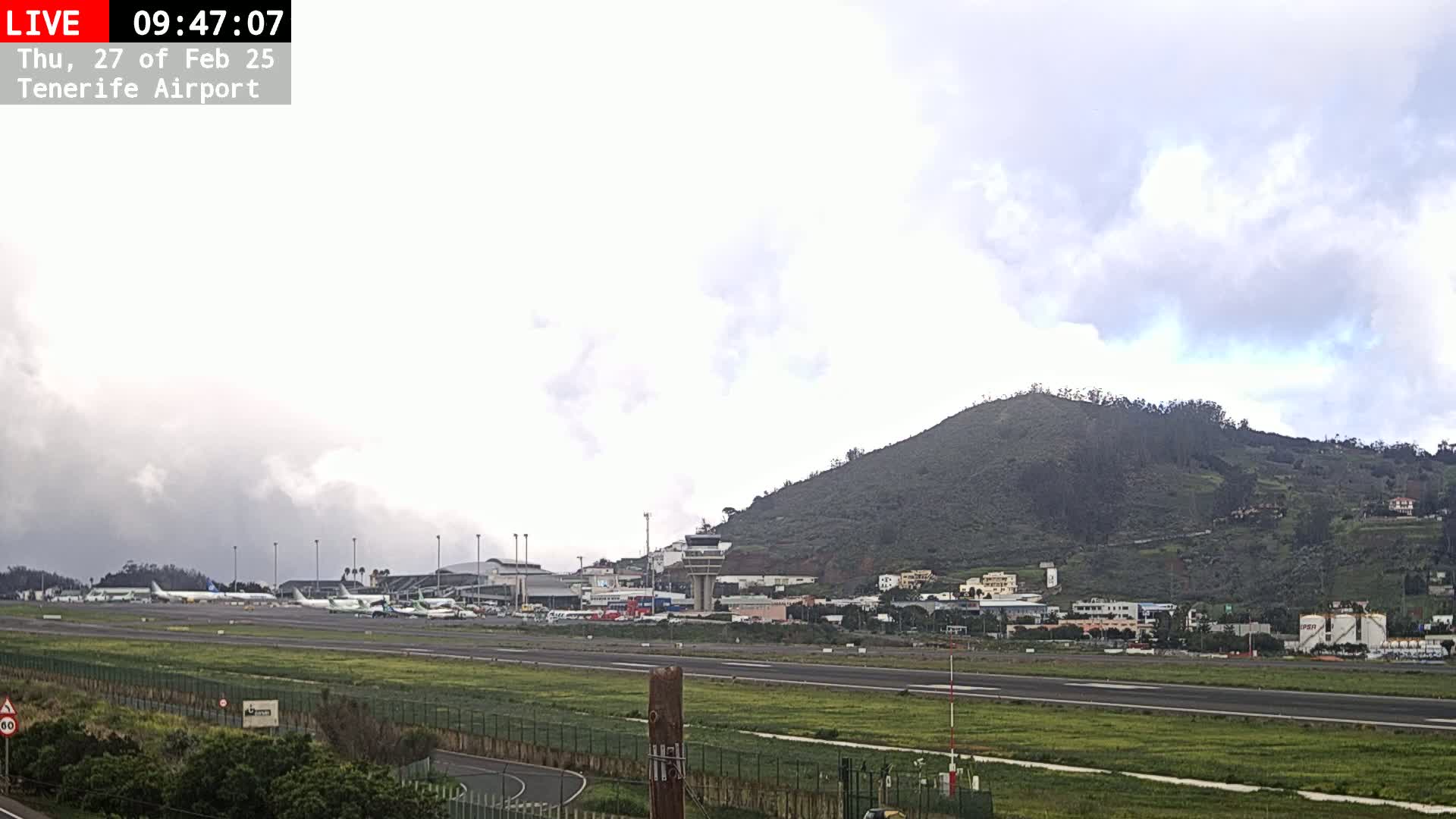 Several airplanes are parked on an airport tarmac next to a grassy area, with a hill and buildings visible in the background under a cloudy sky.