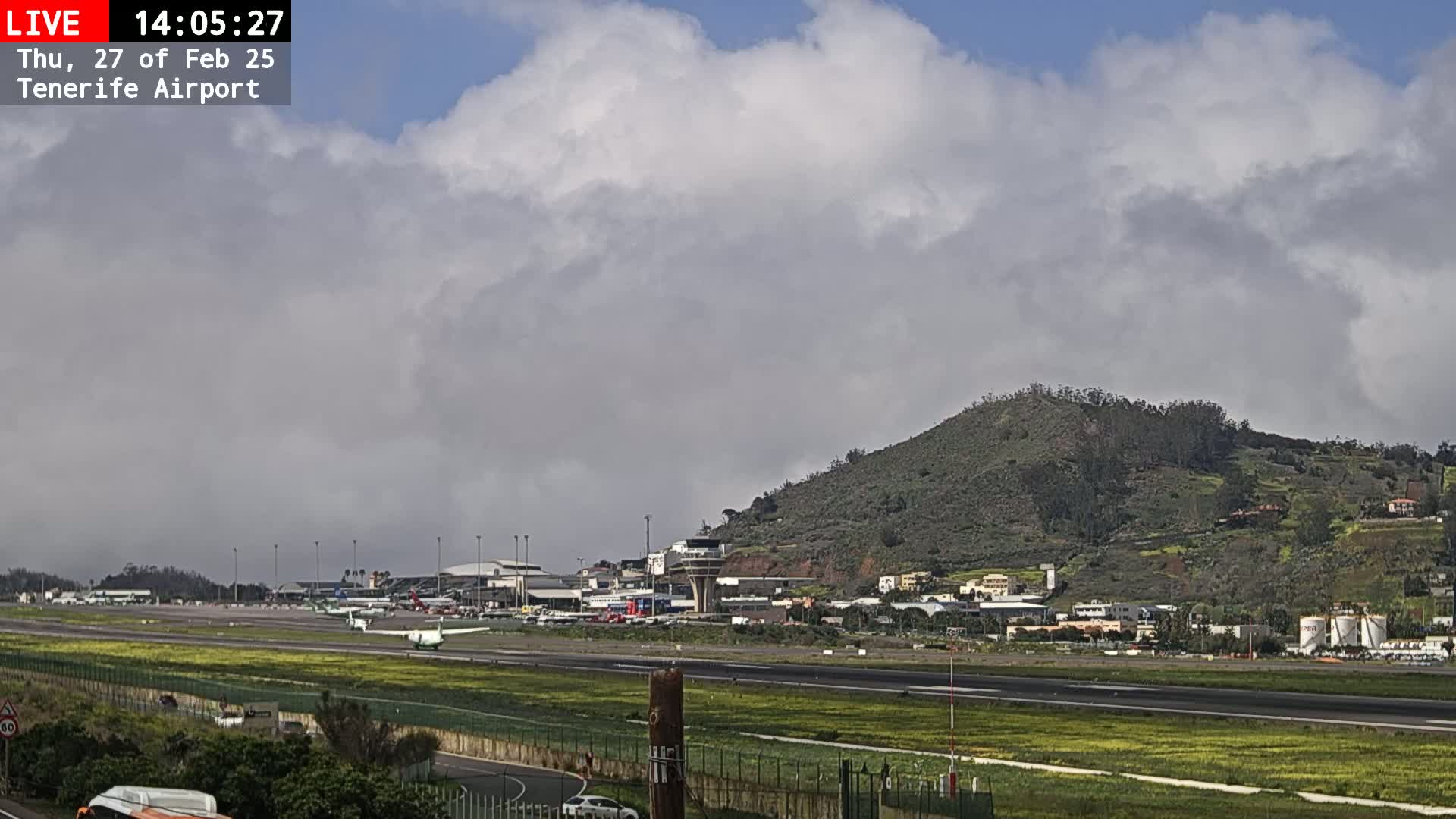 An airport runway with planes, airport buildings, and a green hill in the background under a partly cloudy sky.