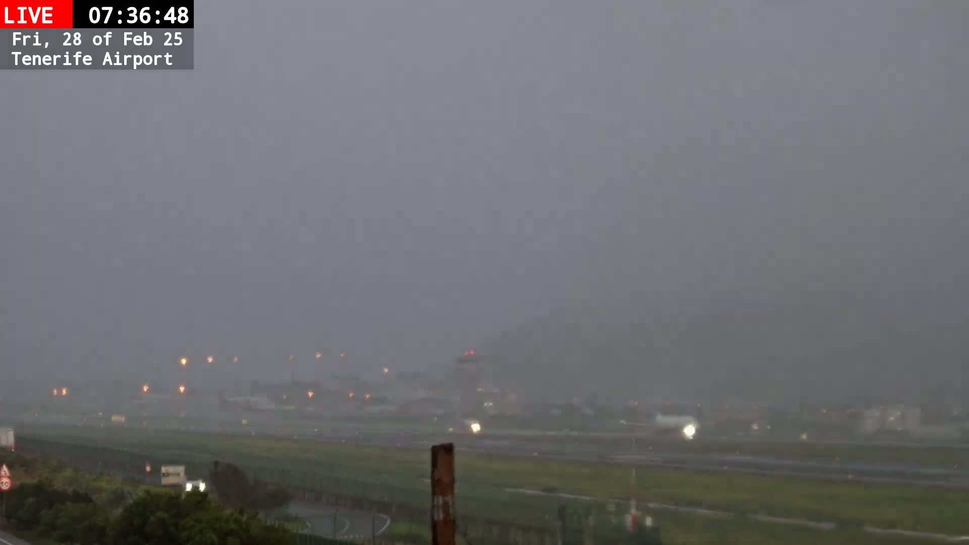 A rain-soaked airport runway is visible through heavy fog, with faint lights illuminating the tarmac and surrounding buildings.