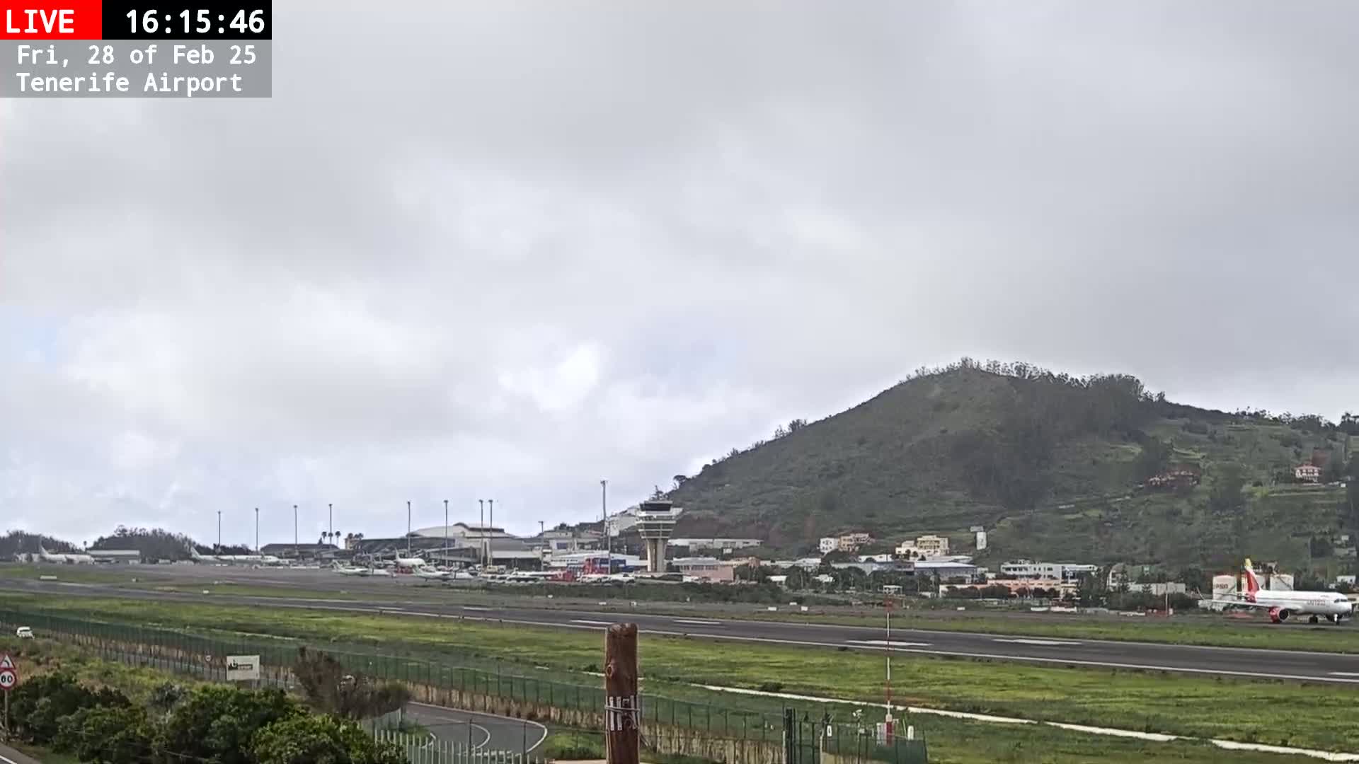 An airport runway with several airplanes, airport buildings, and a green hill in the background under an overcast sky.