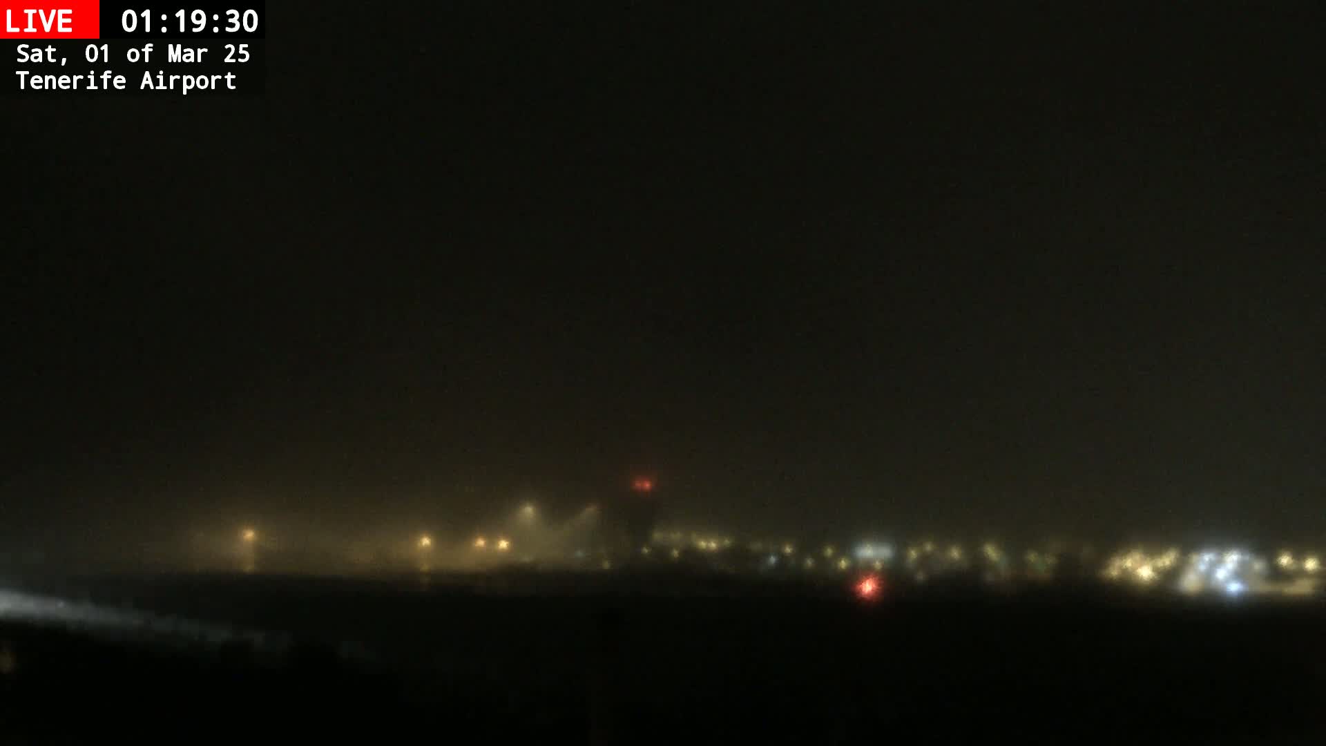 A foggy nighttime view of an airport runway with dimly lit buildings and lights in the distance.