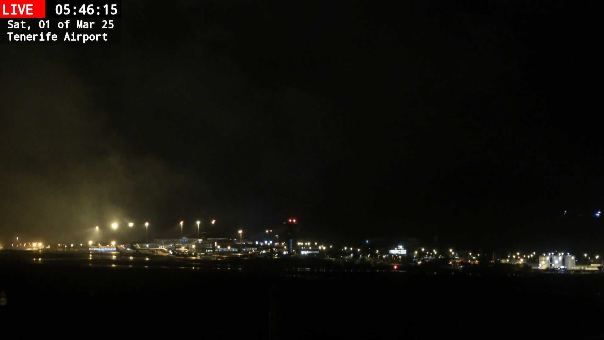A nighttime view of an airport tarmac with several aircraft and buildings faintly visible under a dark, hazy sky.