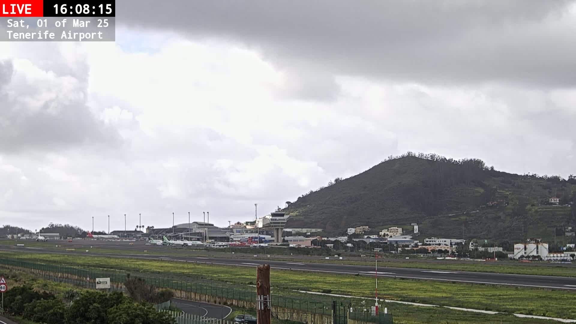An airport runway with several airplanes, surrounded by grassy fields, and a hill in the background under an overcast sky.