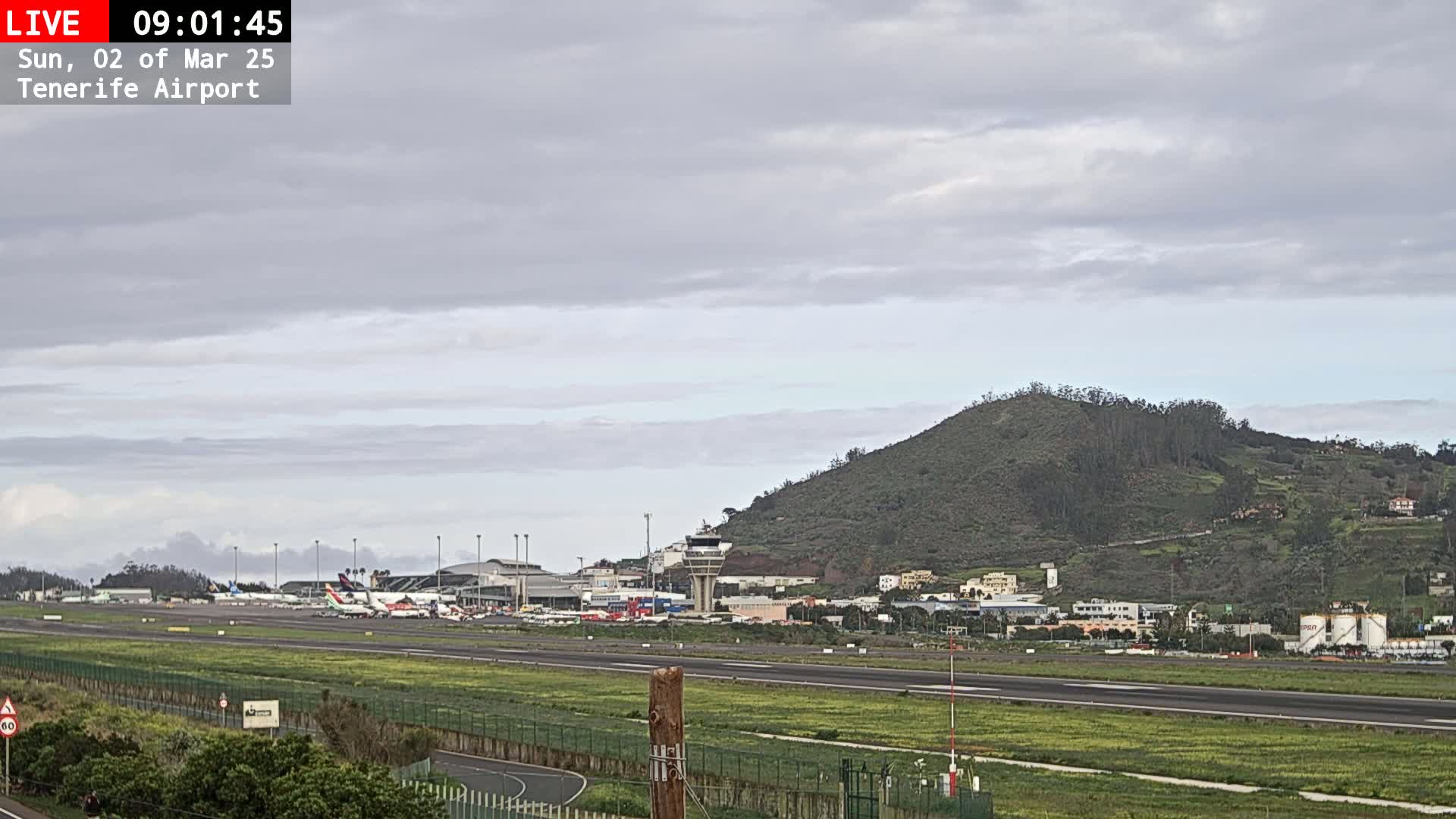 A mostly cloudy day reveals an airport tarmac with several airplanes, surrounded by buildings and a large green hill.