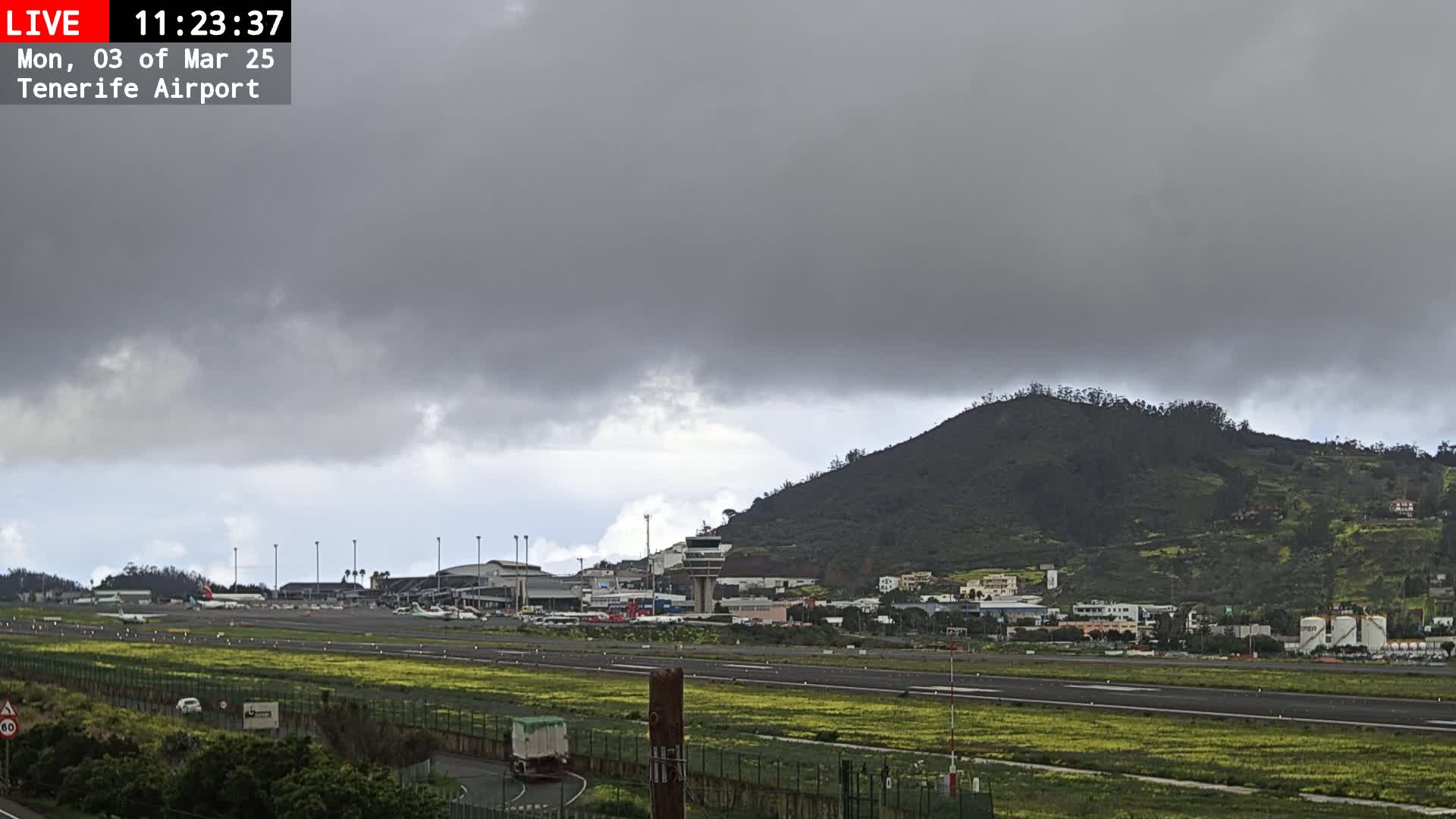 A view of an airport runway, terminal buildings, and a nearby hill under an overcast sky.