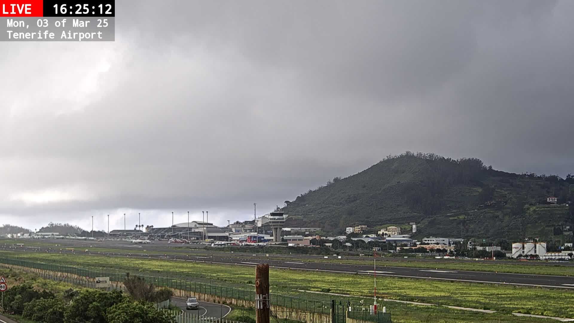 An airport runway and terminal are visible beneath an overcast sky, with a large hill in the background.