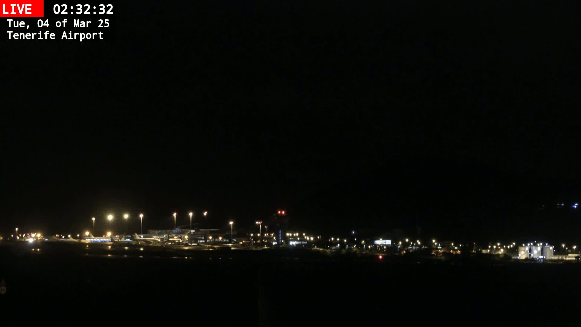 A nighttime, long shot of an airport with several airplanes visible on the tarmac, illuminated by bright lights under a dark sky.