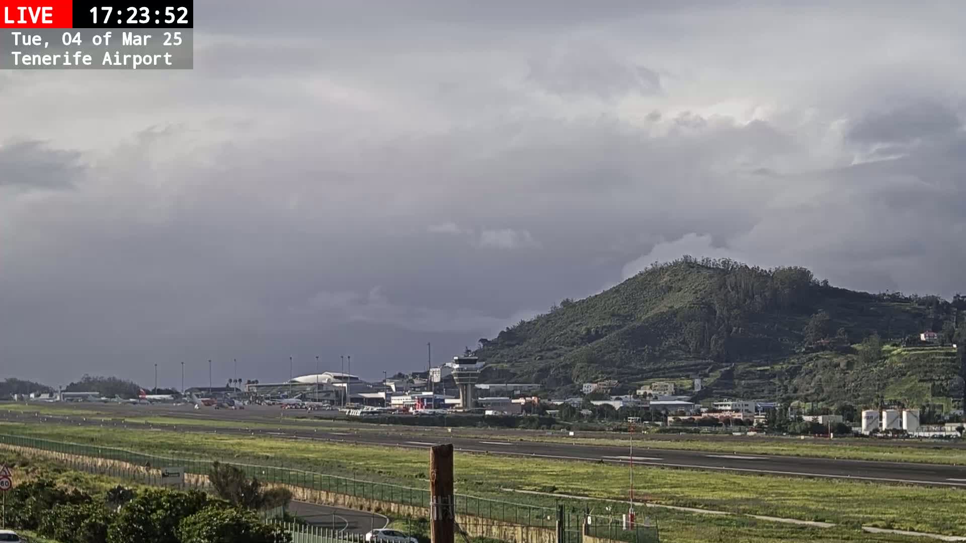 An airport runway and terminal buildings are visible beneath an overcast sky, with a large, green hill in the background.