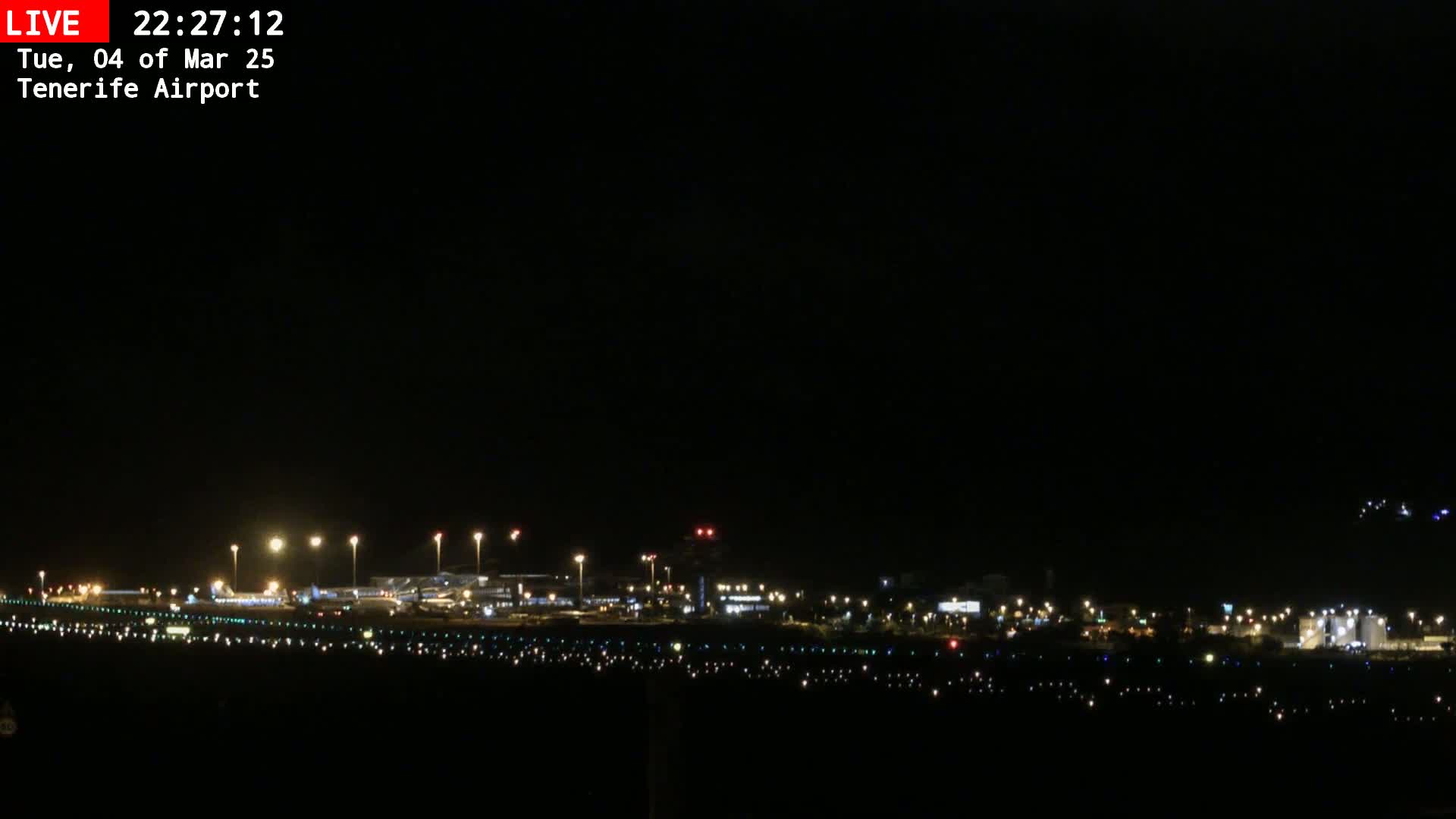 A nighttime long shot of an airport runway and terminal buildings illuminated under a dark sky.