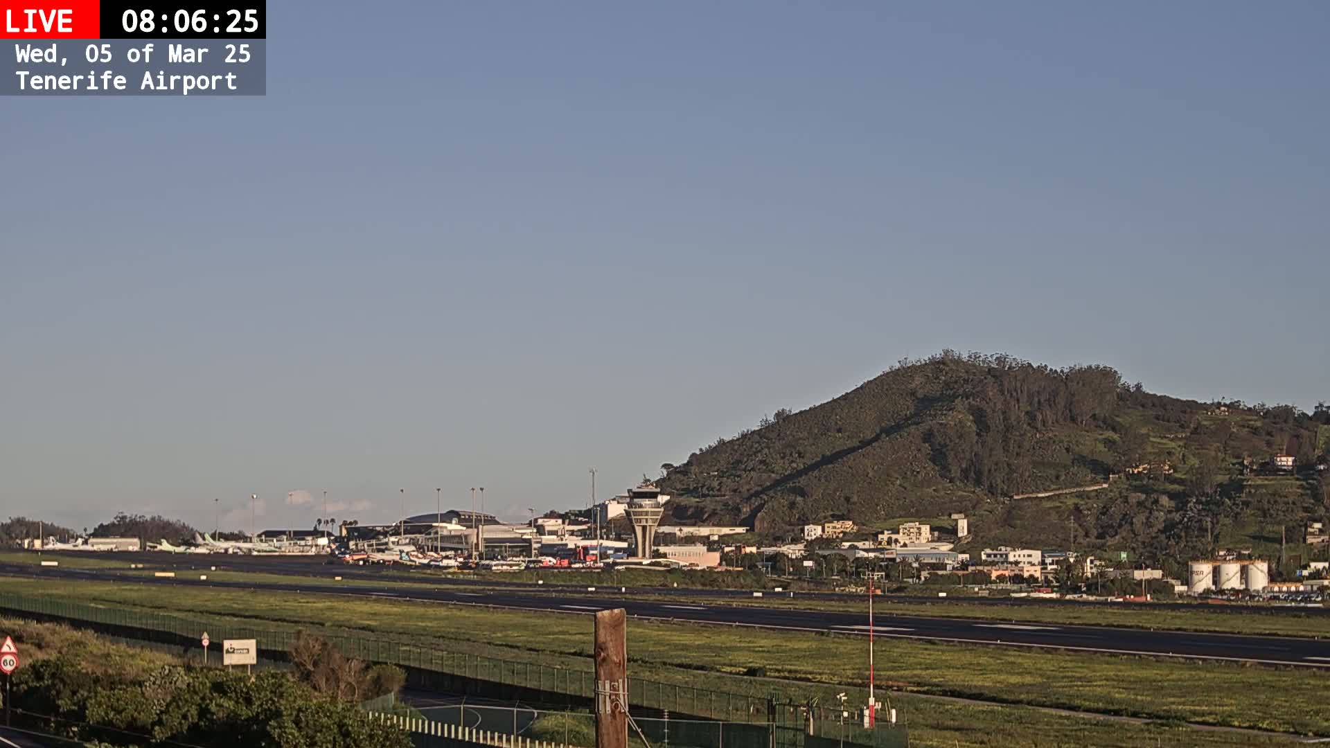 A long view of an airport runway, terminal buildings, and a control tower, set against a clear sky and a verdant hill in the background.