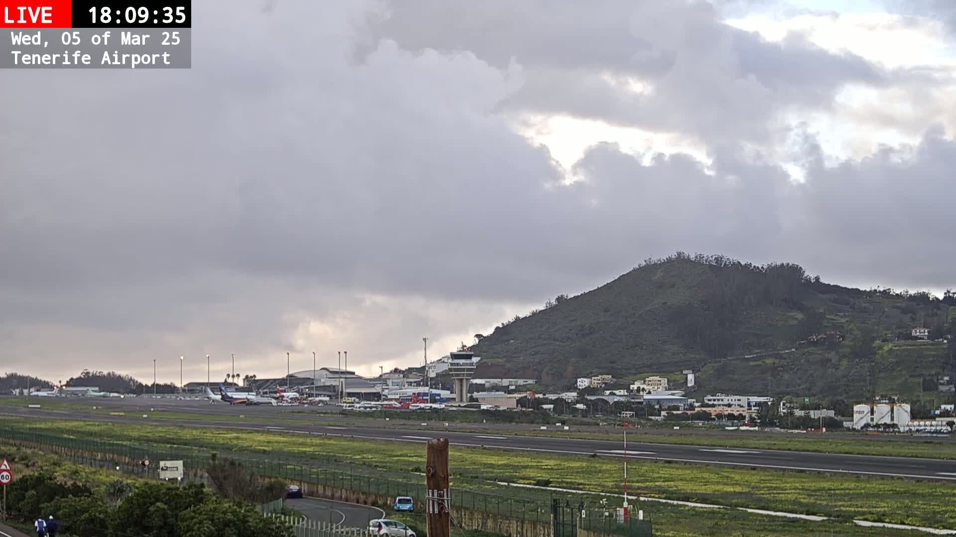 An overcast sky hangs over an airport runway, with several airplanes parked near the terminal buildings and a hill in the background.