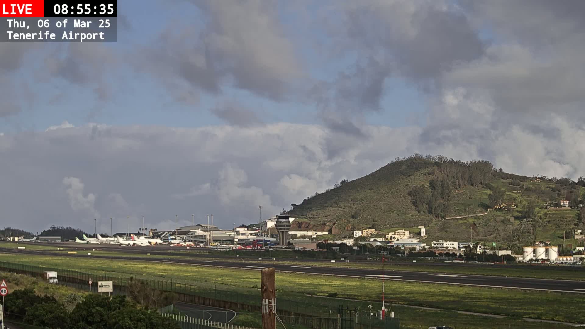 A partly cloudy sky overlooks an airport with several airplanes parked on the tarmac, a control tower, and a green hill in the background.