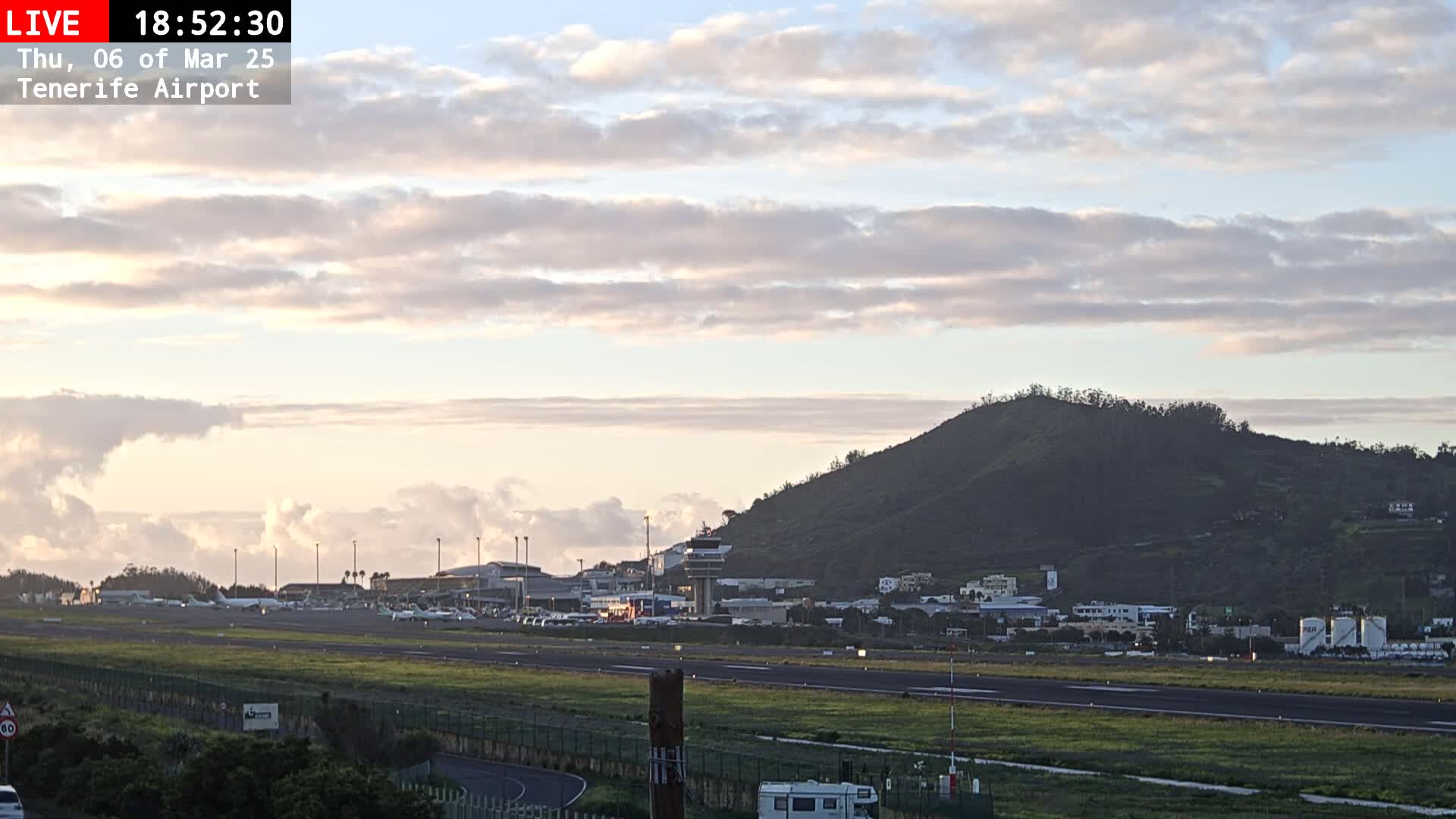 A mostly cloudy sunset illuminates an airport runway with several airplanes parked, a control tower, and a large hill in the background.
