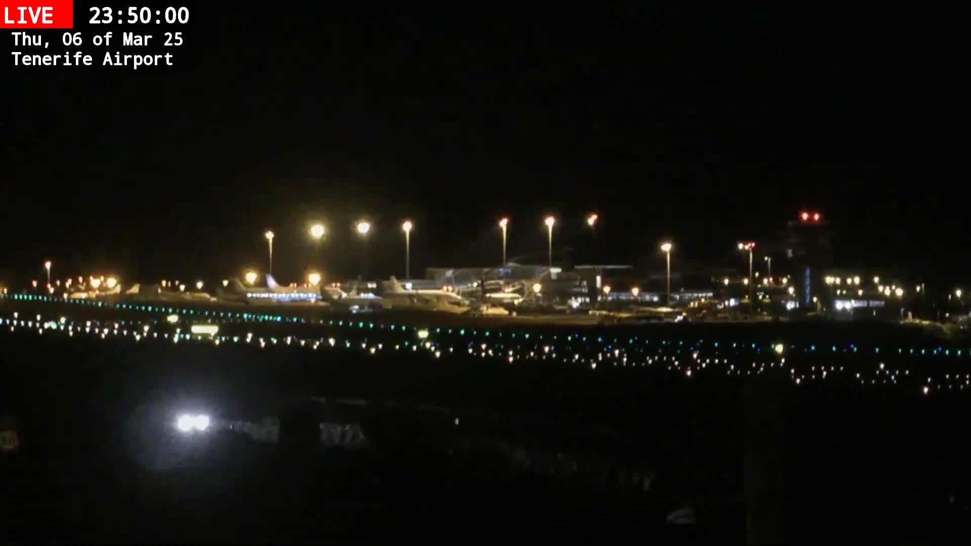 A nighttime view of a brightly lit airport with several airplanes parked on the tarmac under a clear, dark sky.