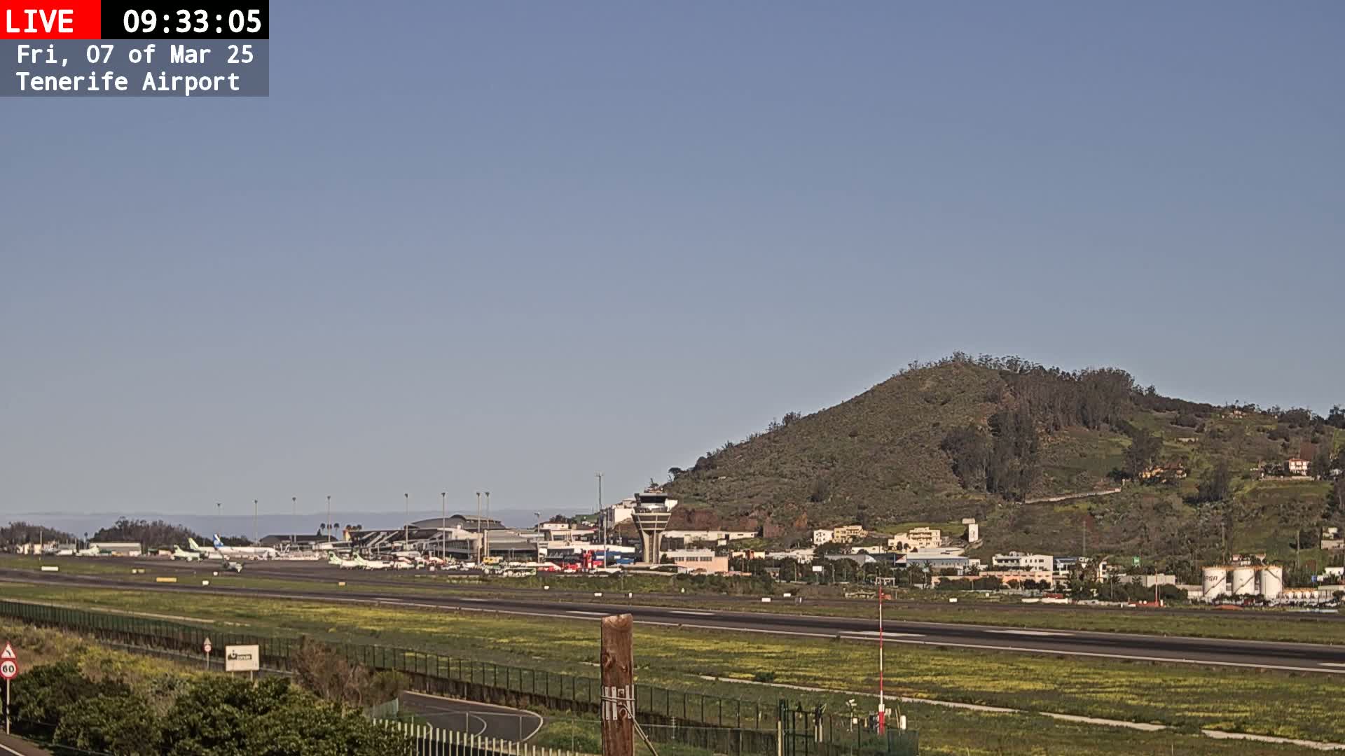 An airport runway and terminal are visible beneath a clear sky, with a hilly landscape in the background.