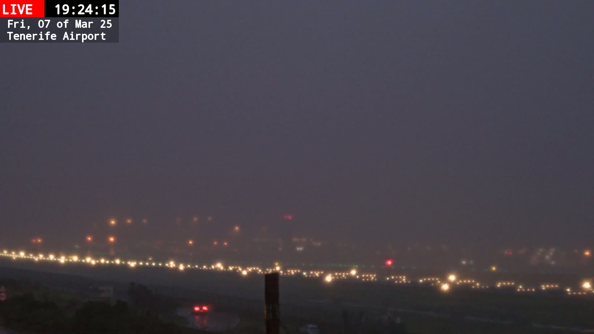 A foggy night scene shows a line of runway lights at an airport, with a few vehicles visible in the foreground.