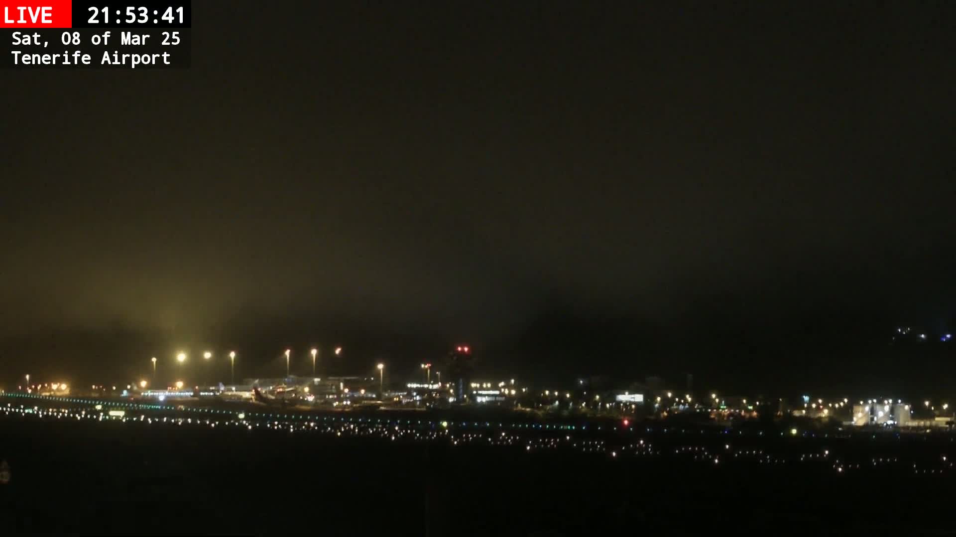 A nighttime, foggy view of an airport runway and terminal buildings with lights illuminating the scene.