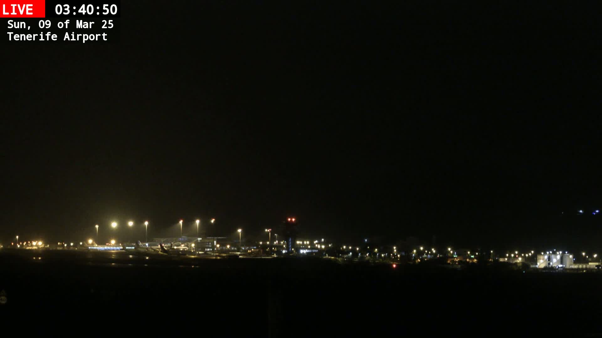 A nighttime view of an airport runway and terminal buildings under a dark, possibly overcast sky.