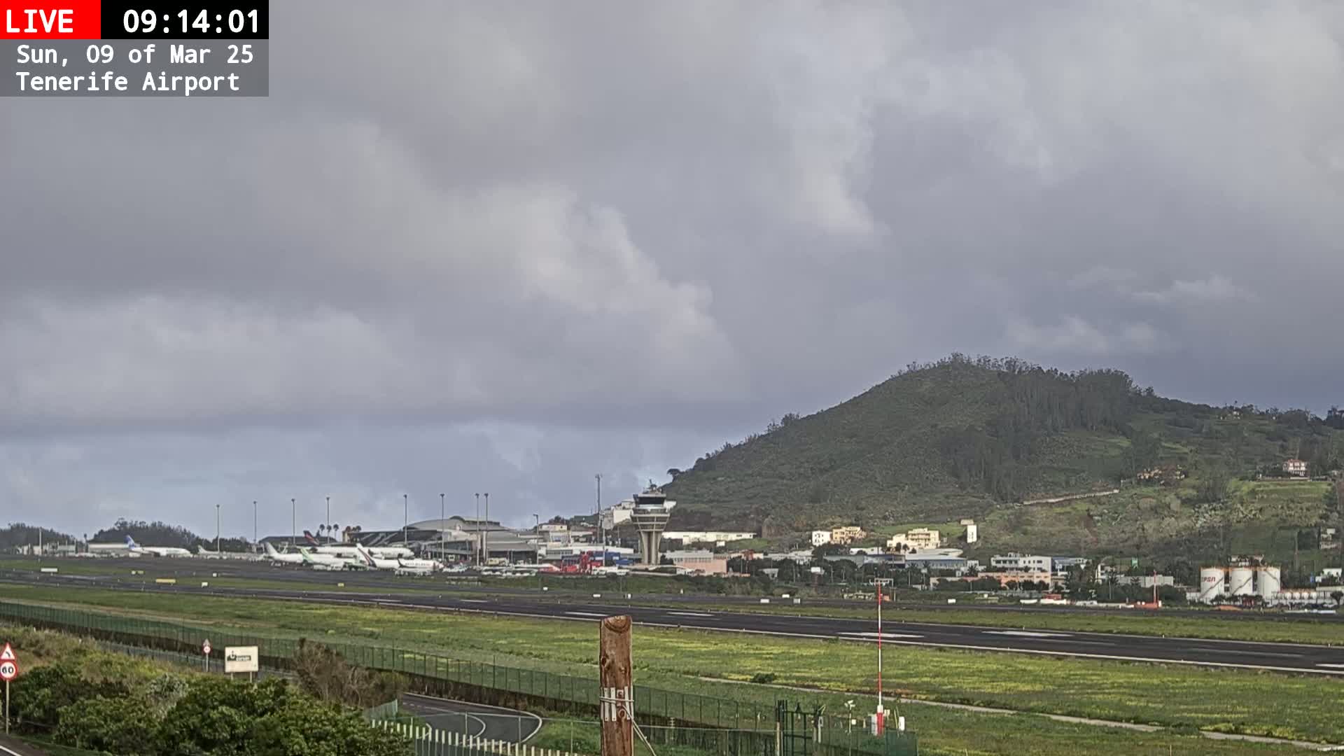 An airport tarmac with several airplanes, airport buildings, and a green hill in the background under an overcast sky.