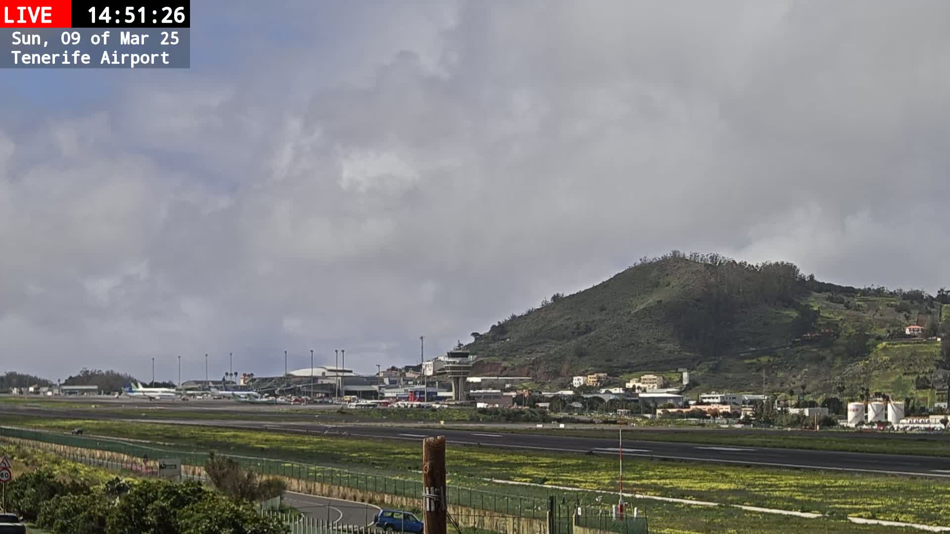 An airport runway and terminal are visible beneath a mostly cloudy sky, with a green hill in the background.