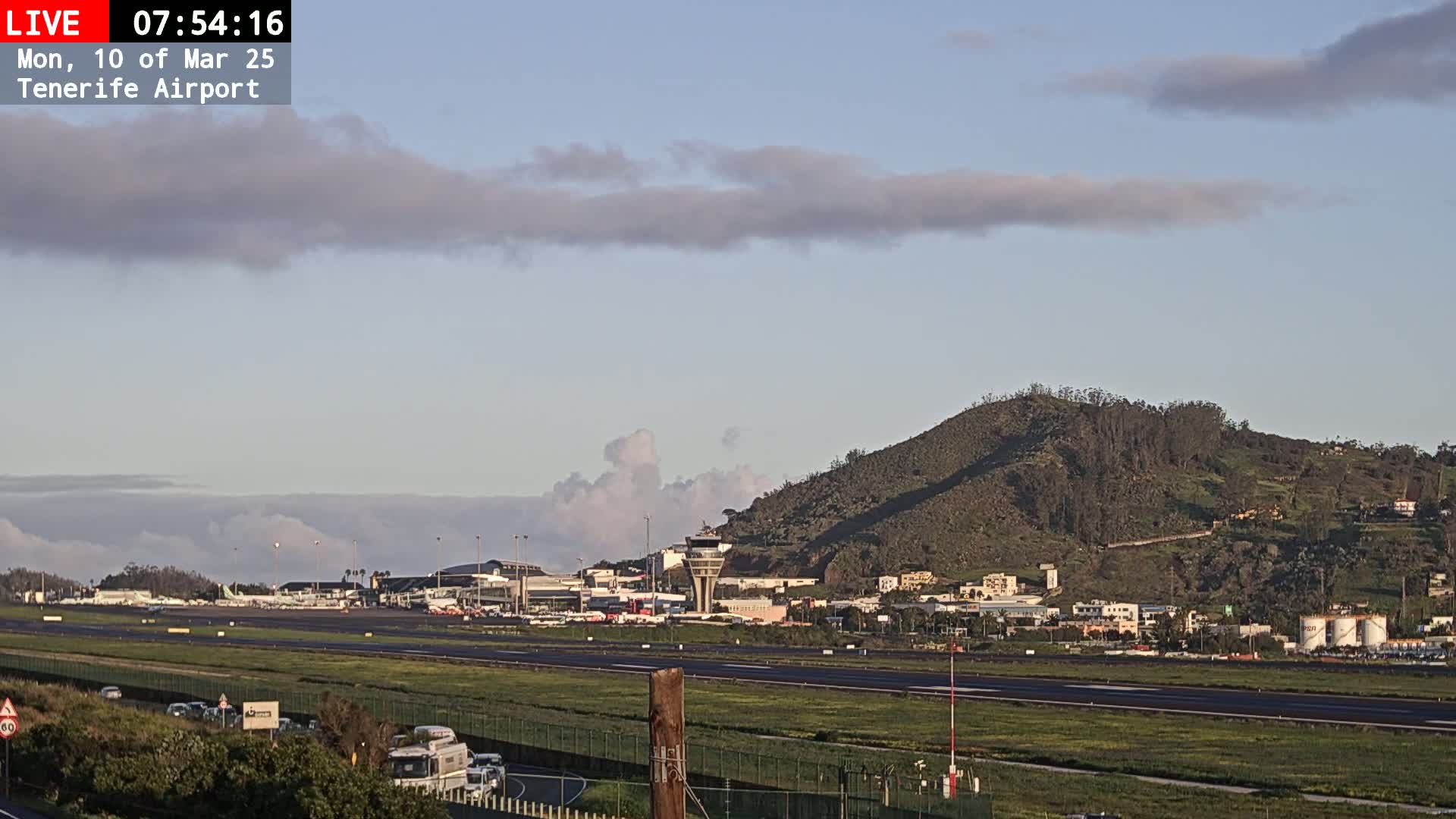A partly cloudy sky overlooks an airport runway with planes parked at the terminal, a control tower, and a hill in the background.