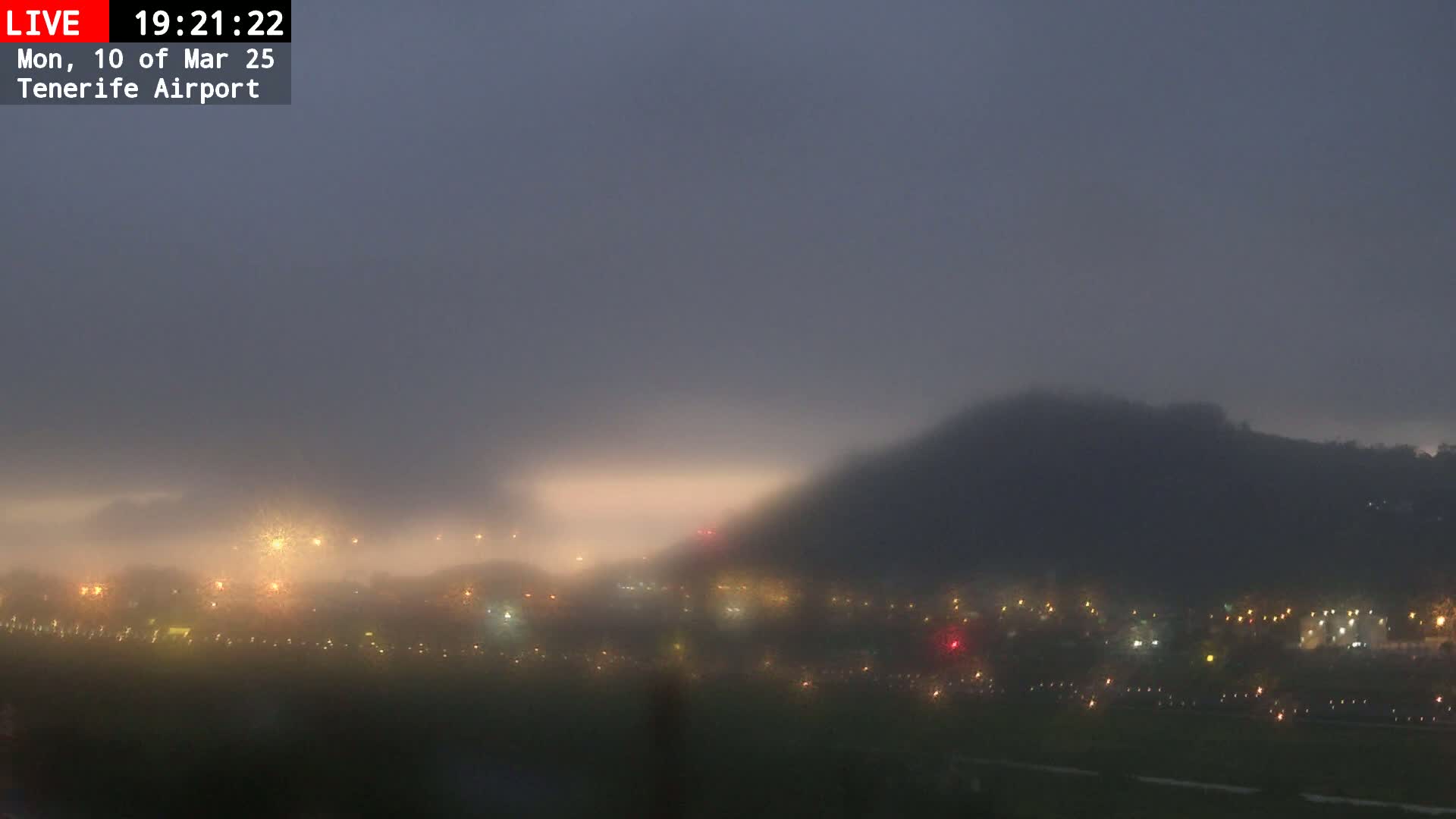 A foggy, dimly lit landscape at night shows a hill in the background and a scattering of lights in the foreground.