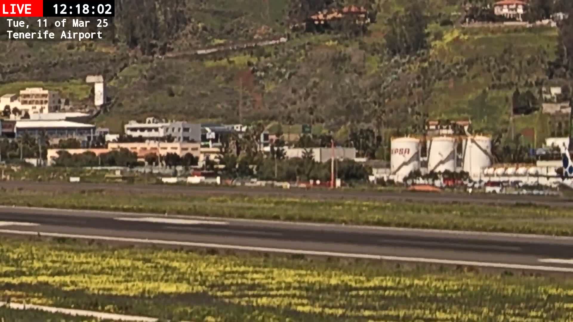 An airport runway with buildings and fuel tanks in the background on a sunny day.