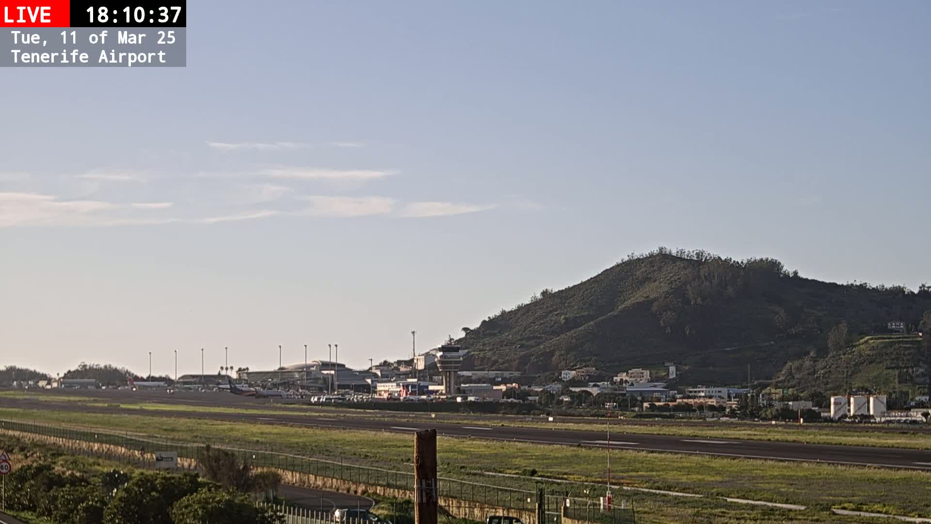 A runway at an airport is shown under a mostly clear sky with a few wispy clouds, with a large hill visible in the background.