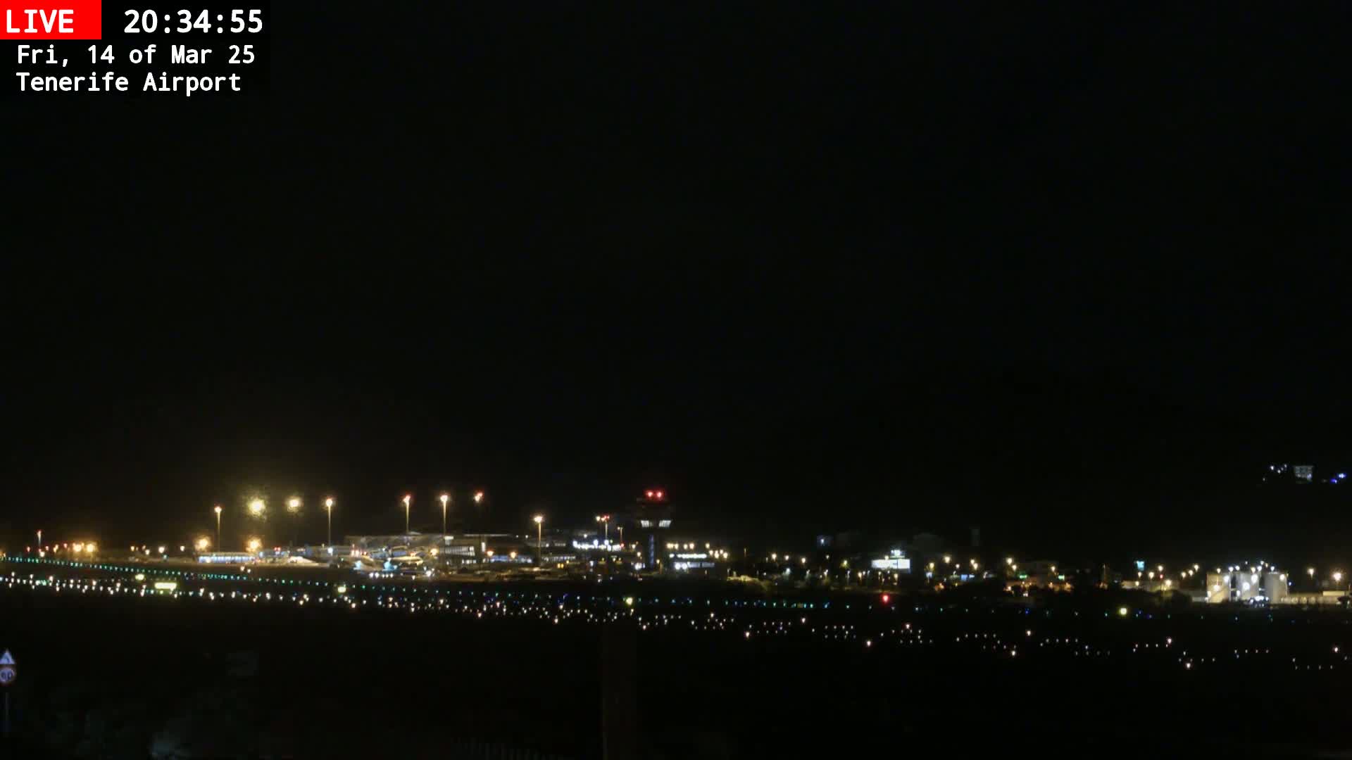 A nighttime long shot of a brightly lit airport runway and terminal buildings under a clear, dark sky.