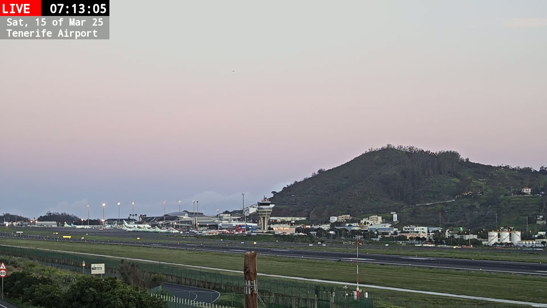 Under a pale pink dawn sky, an airport with numerous airplanes parked on the tarmac is situated in front of a large, green hill.