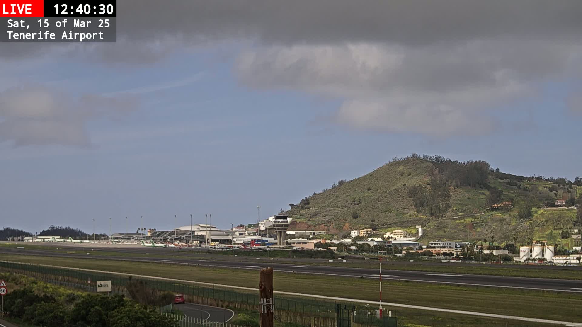 A partly cloudy sky overlooks an airport runway with several planes parked, surrounded by buildings and a green hill.