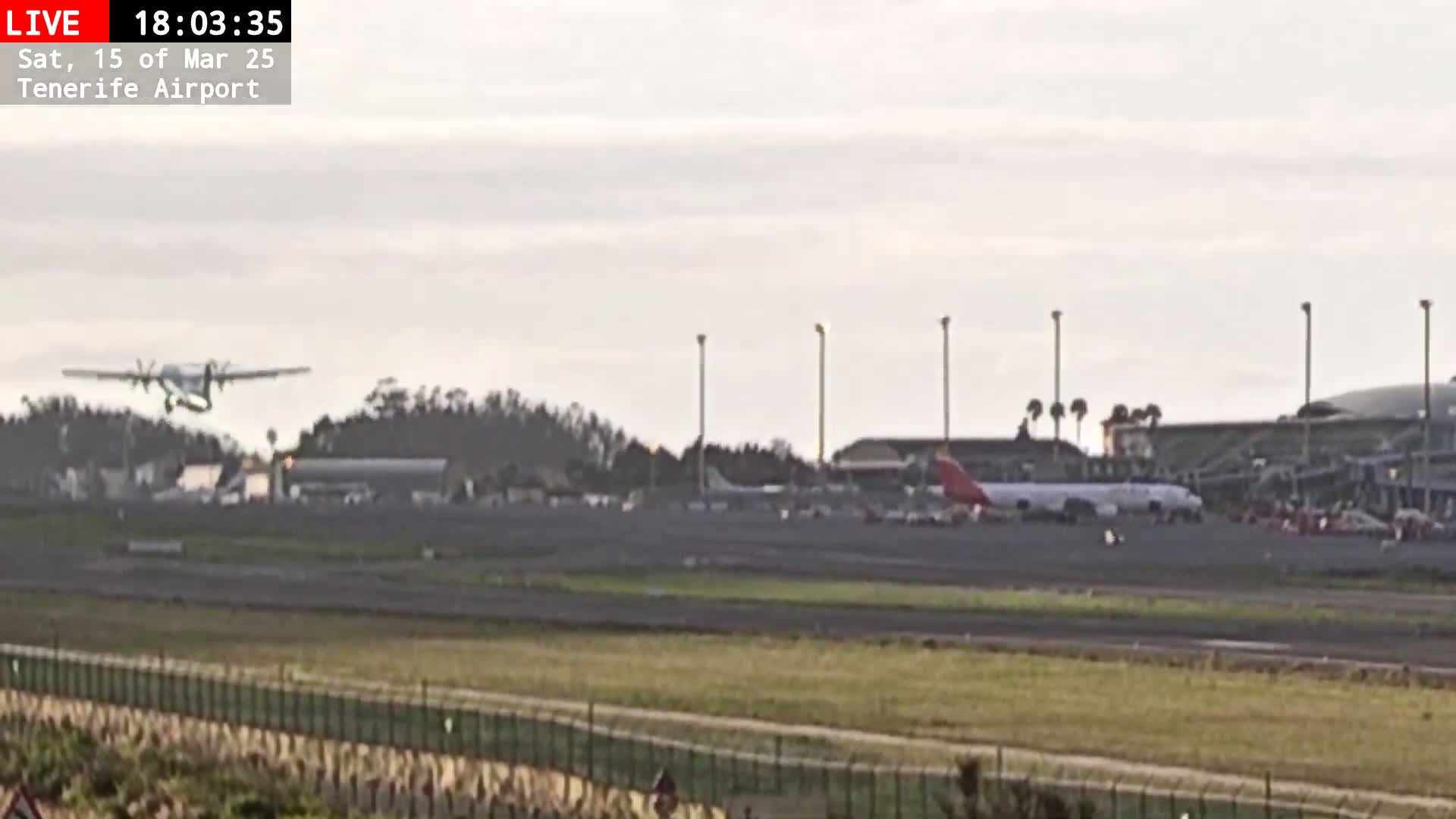 A small airplane is taking off from an airport runway on an overcast day, with a larger airplane parked on the tarmac in the background.