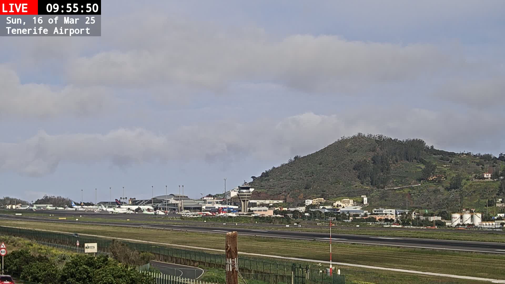 A partly cloudy day at Tenerife Airport shows numerous airplanes parked on the tarmac, with a green hill in the background.